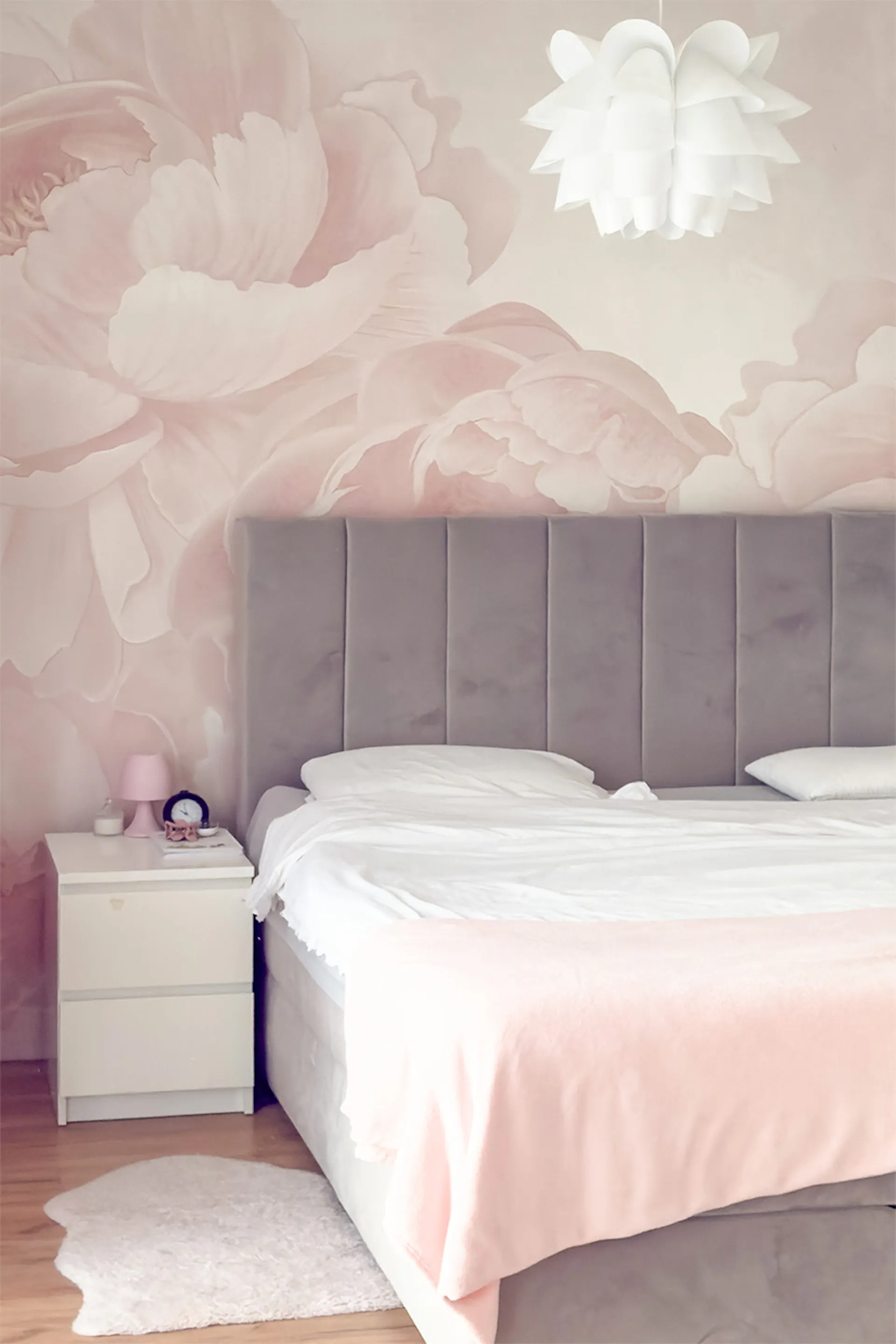 Łóżko z szarym pikowanym wezgłowiem; Fototapeta z dużymi różowymi kwiatami w sypialni; Biała szafka nocna.