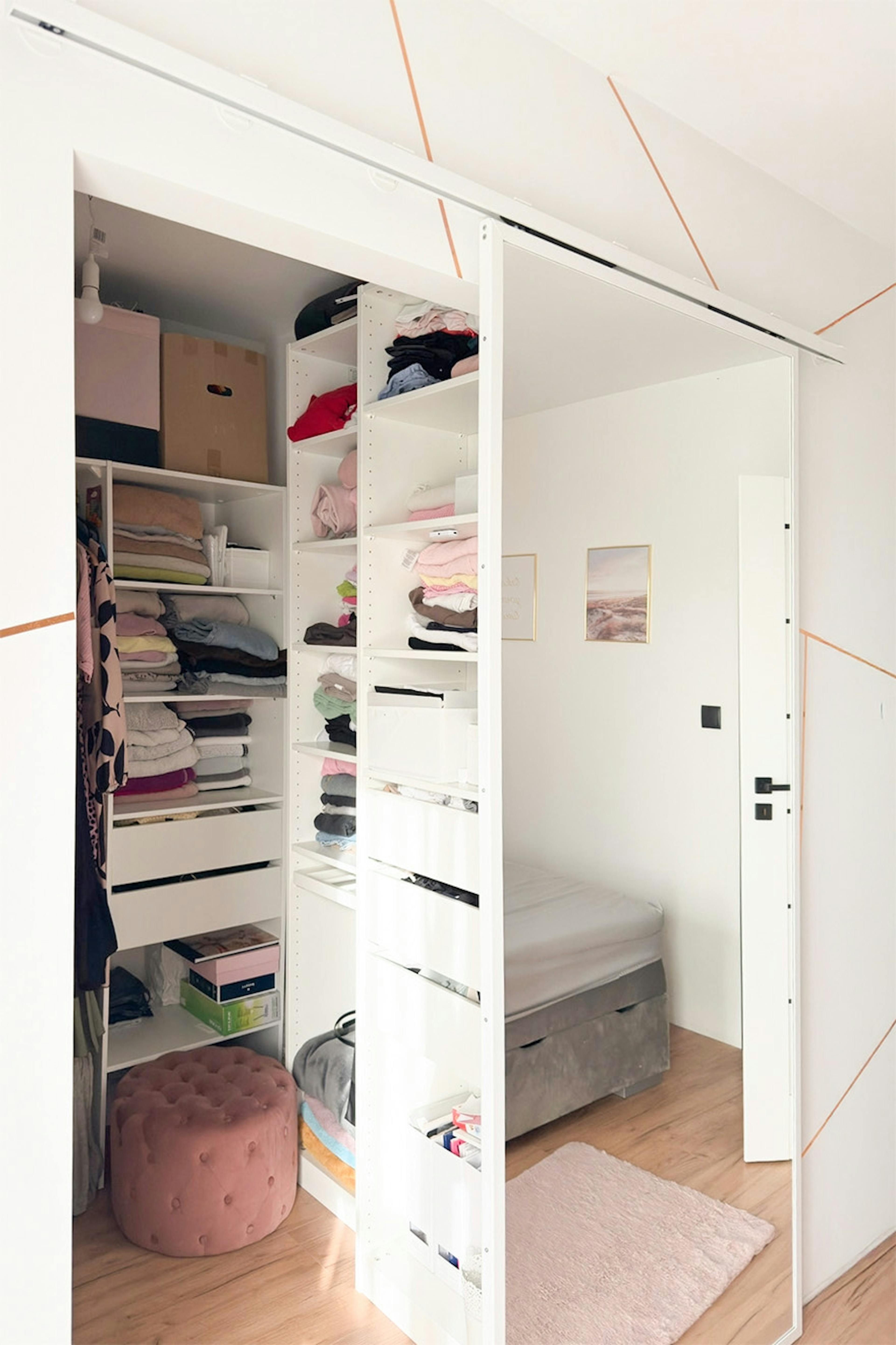 Offenes, weißes Ankleidezimmer mit Regalen und Schubladen; Blick auf das Bett, das sich im Spiegel widerspiegelt; Pudertöniger Velours-Hocker im Kleiderschrank.