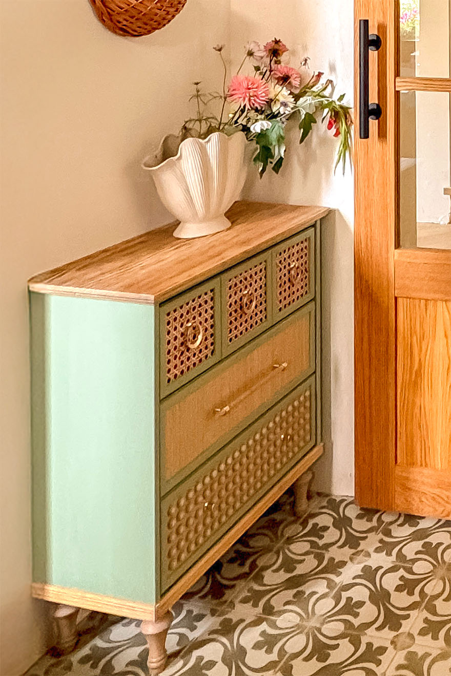 Szałwiowa komoda z rattanowymi frontami i złotymi uchwytami stojąca w wiatrołapie.