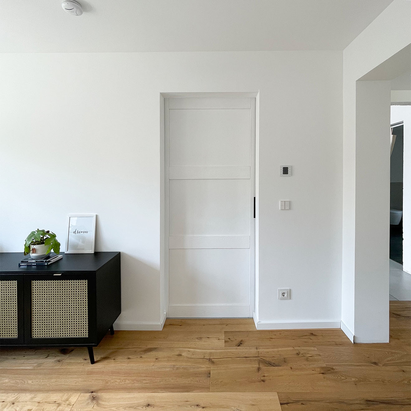 Schwarze niedrige Kommode mit Rattan auf weißem Wandhintergrund, weiße Massivholz-Schiebetür, weiße Tür auf weißer Wand