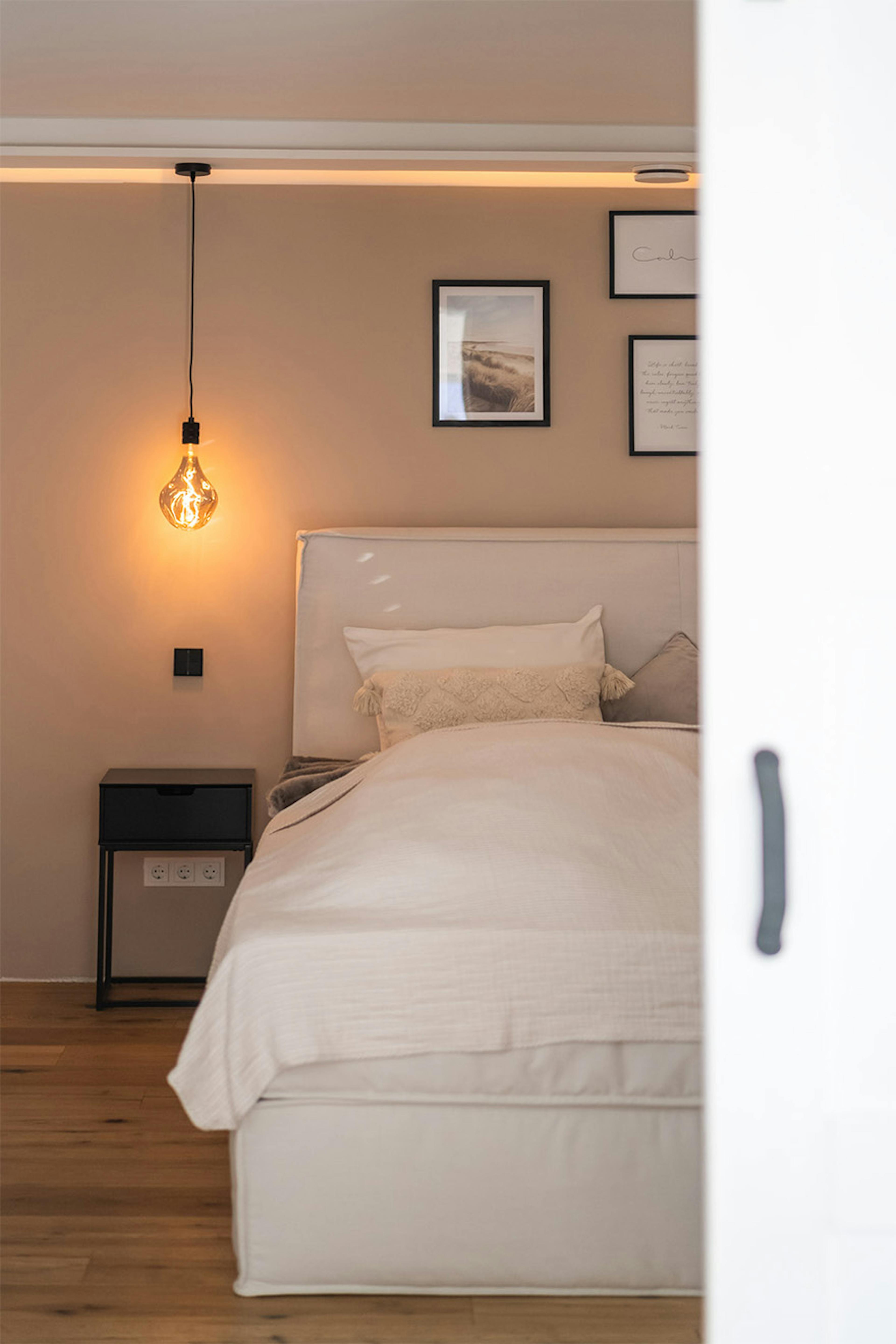 hängende Glühbirnenlampe, ein kleiner schwarzer Nachttisch, ein Bett in einer beigen Schlafzimmeranordnung