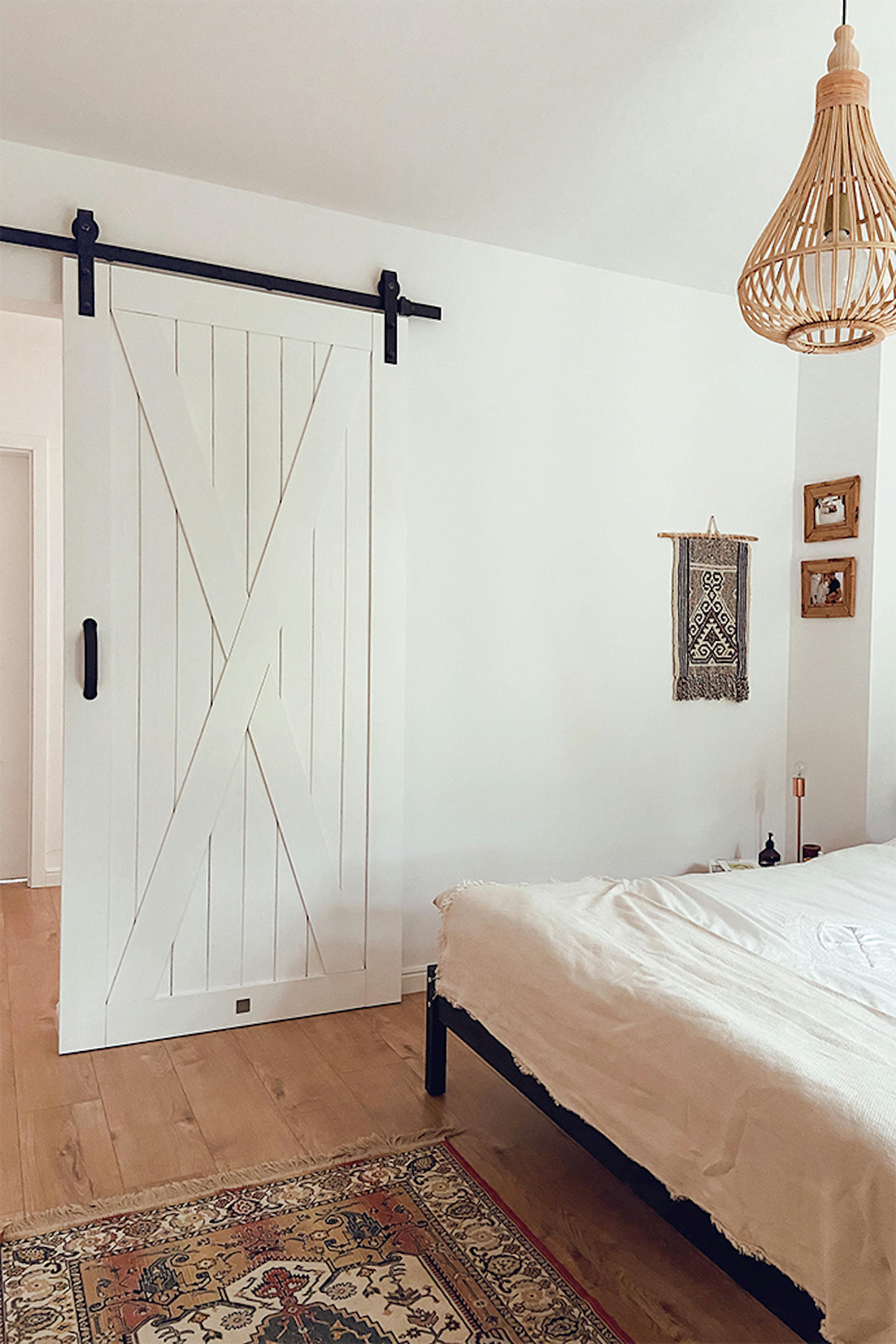 Weiße Massivholz-Schiebetür im Eingang zum Naturschlafzimmer, bunter Teppich vor dem Bett im Schlafzimmer, weiße Wände im Schlafzimmer, Bambus-Pendelleuchte, natürliche Boho-Dekorationen an der weißen Wand