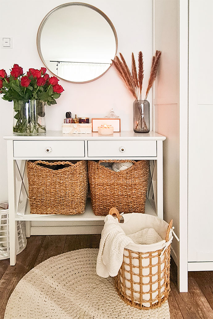 weiße Kommode mit Weidenkörben für Bettwäsche in einem skandinavischen Interieur, lebende Blumendekoration im Schlafzimmer, weiße Wände und weiße Möbel im Schlafzimmer, geflochtener Teppich