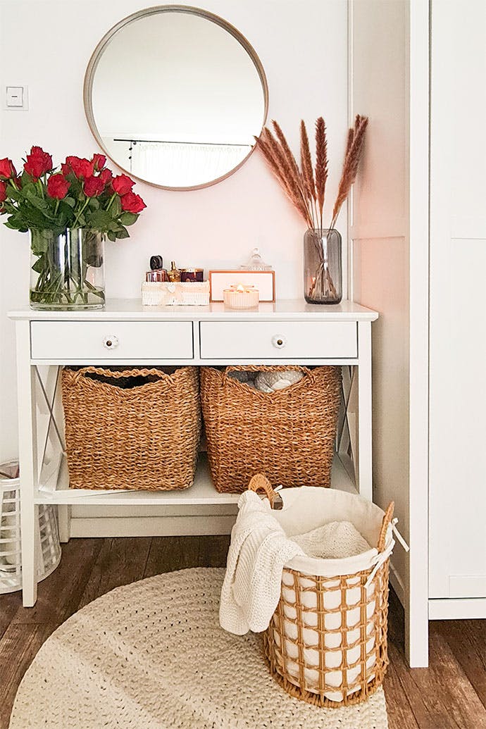 weiße Kommode mit Weidenkörben für Bettwäsche in einem skandinavischen Interieur, lebende Blumendekoration im Schlafzimmer, weiße Wände und weiße Möbel im Schlafzimmer, geflochtener Teppich