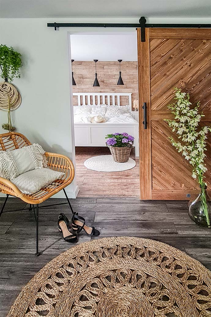 Holzschiebetür im Schlafzimmereingang, Korbsessel mit dekorativen Kissen, geflochtener runder Juteteppich, Blumenschmuck in einem Weidenkorb, weißes Bett in einem Schlafzimmer in skandinavischem Interieur, schwarze Pendelleuchten über einem weißen Bett