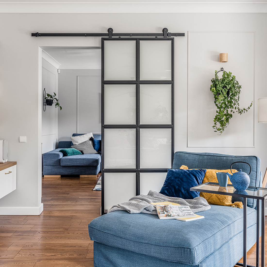 Leseecke, blaues Denim Sofa, Metall- und Glasschiebetür, Industrietür, heller Raum, blaue und gelbe dekorative Kissen, Wandblumenbeet  