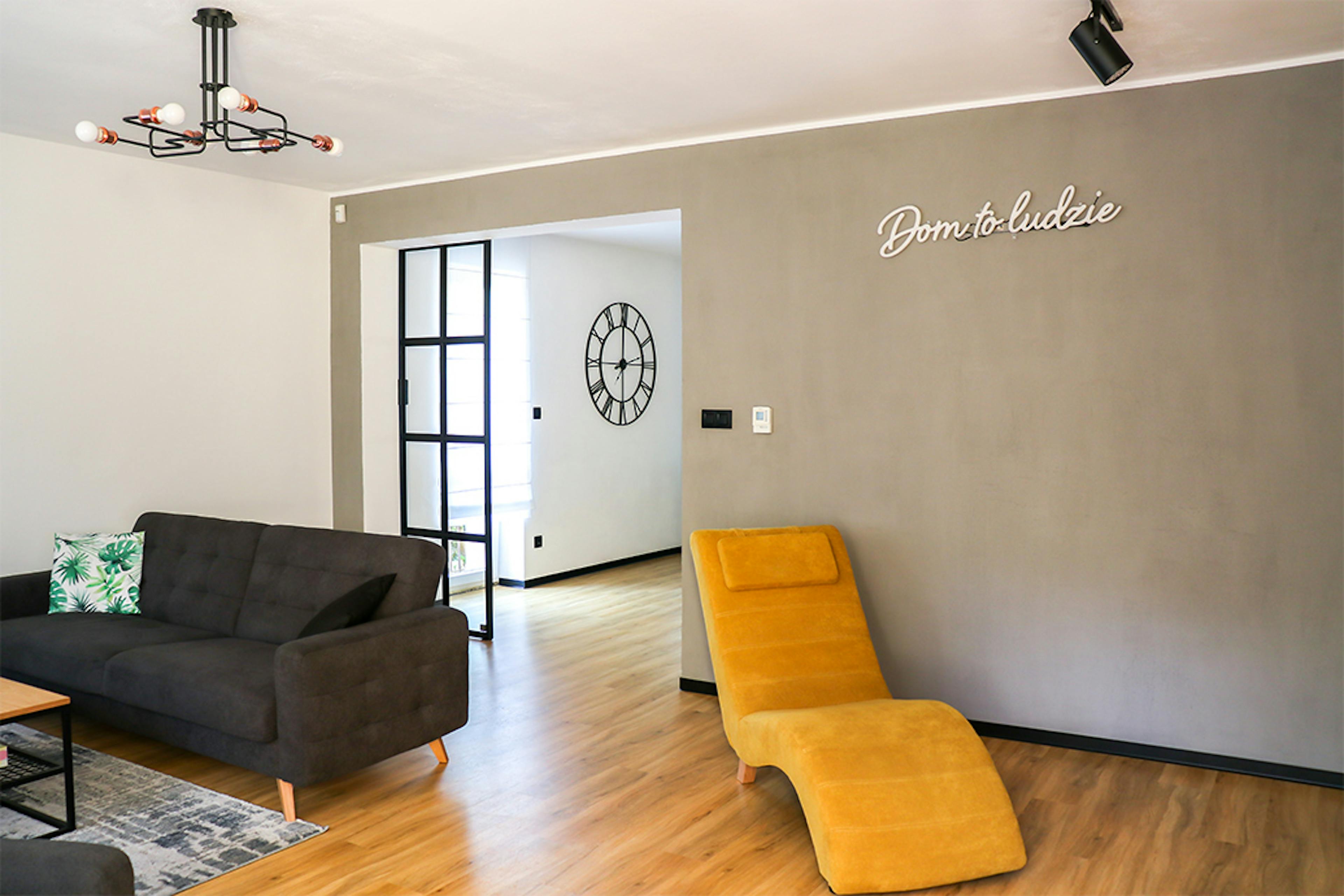 otwarty salon, szara sofa w salonie, industrialna lampa w salonie, żółty fotel do salonu, szara ściana w salonie, salon otwarty na jadalnię, metalowe drzwi ze szkłem, drzwi przesuwne ze szkłem, drzwi w stylu loft, neonowy napis na ścianę, neonowe dekoracje