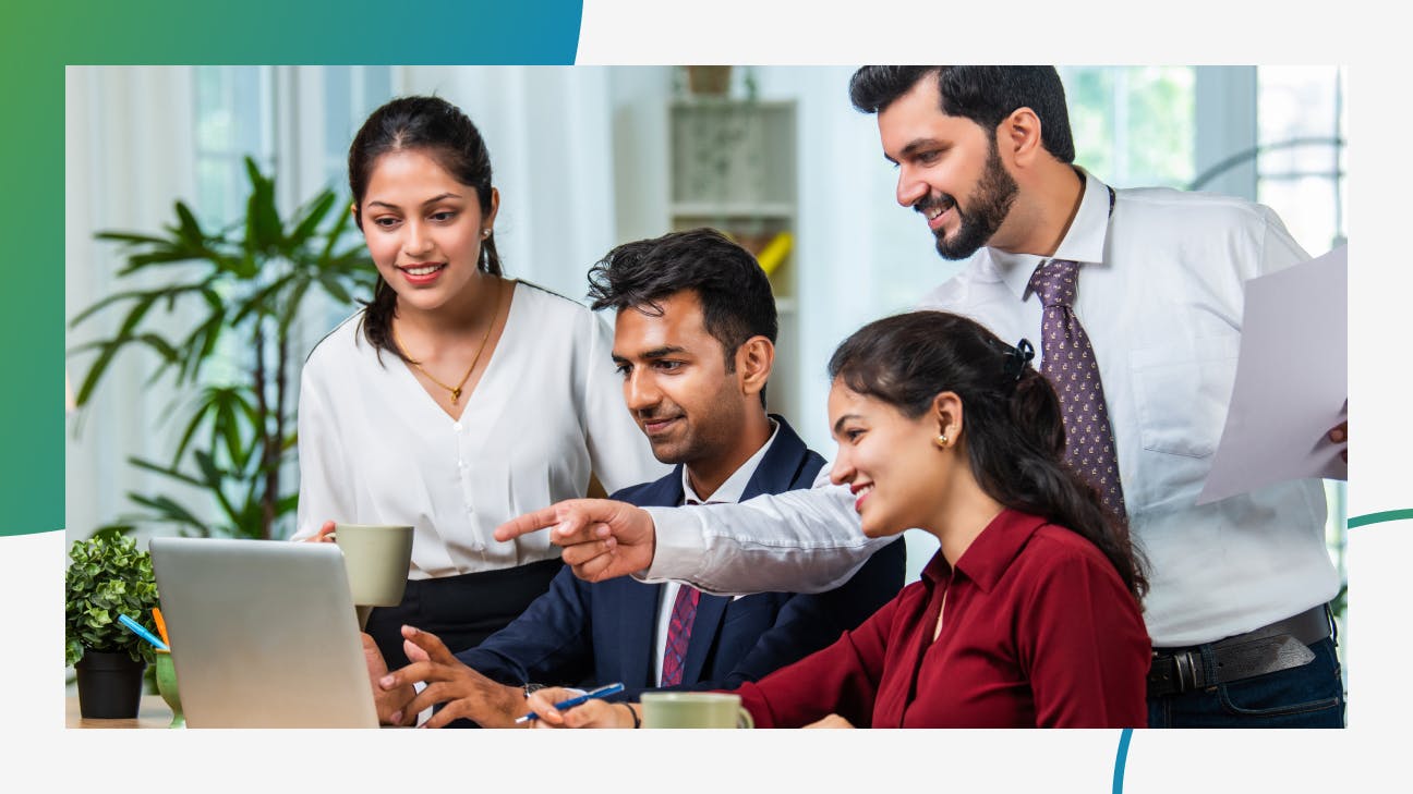 Four people standing and sitting around a laptop in an office environment
