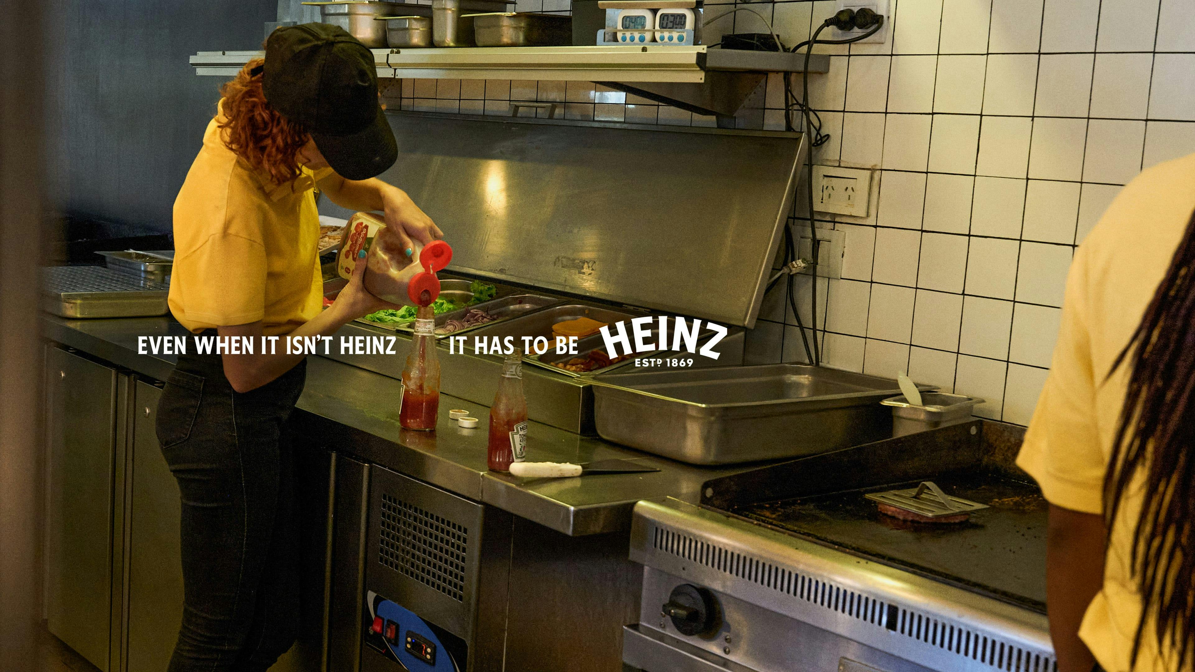 Une personne portant un chandail jaune remplit une bouteille de ketchup Heinz à partir d’une bouteille d’une autre marque de ketchup dans une cuisine de restaurant. 