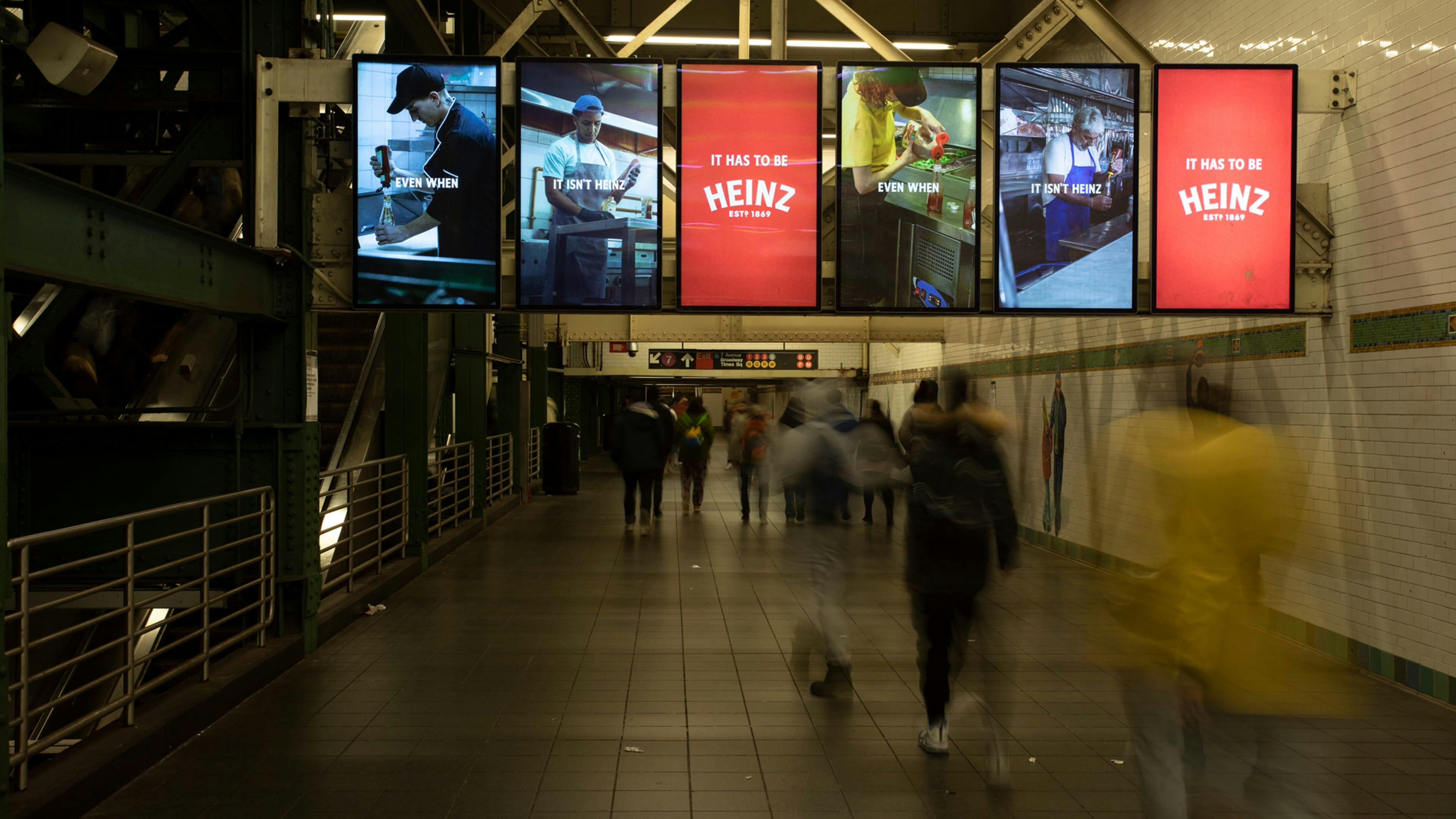 A subway station with several digital advertisements aligned overhead, each showing a different person pouring non-Heinz ketchup into Heinz ketchup bottles, with the tagline "Even when it isn't Heinz, it has to be Heinz."