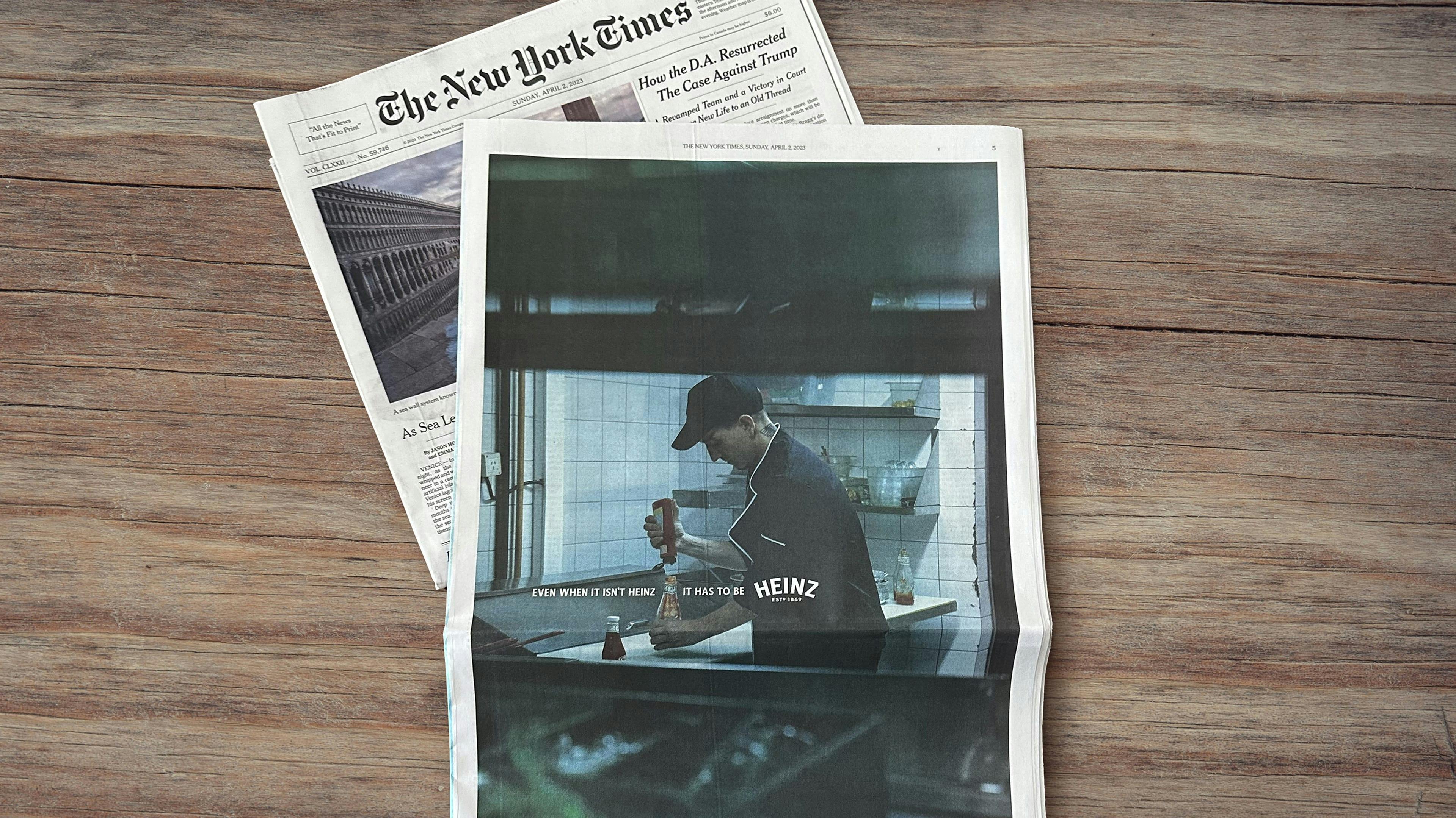 The New York Times newspaper lies on a wooden surface, featuring an advertisement with a chef squeezing non-Heinz ketchup into a Heinz ketchup bottle and the tagline “Even when it isn’t Heinz, it has to be Heinz.”