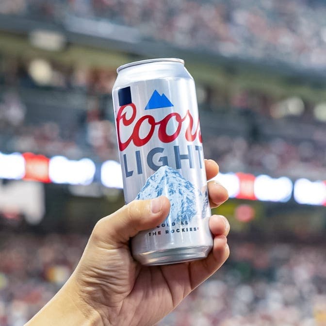 A hand holding a special-edition Coors Light can with a black square on the label, in a baseball stadium. 