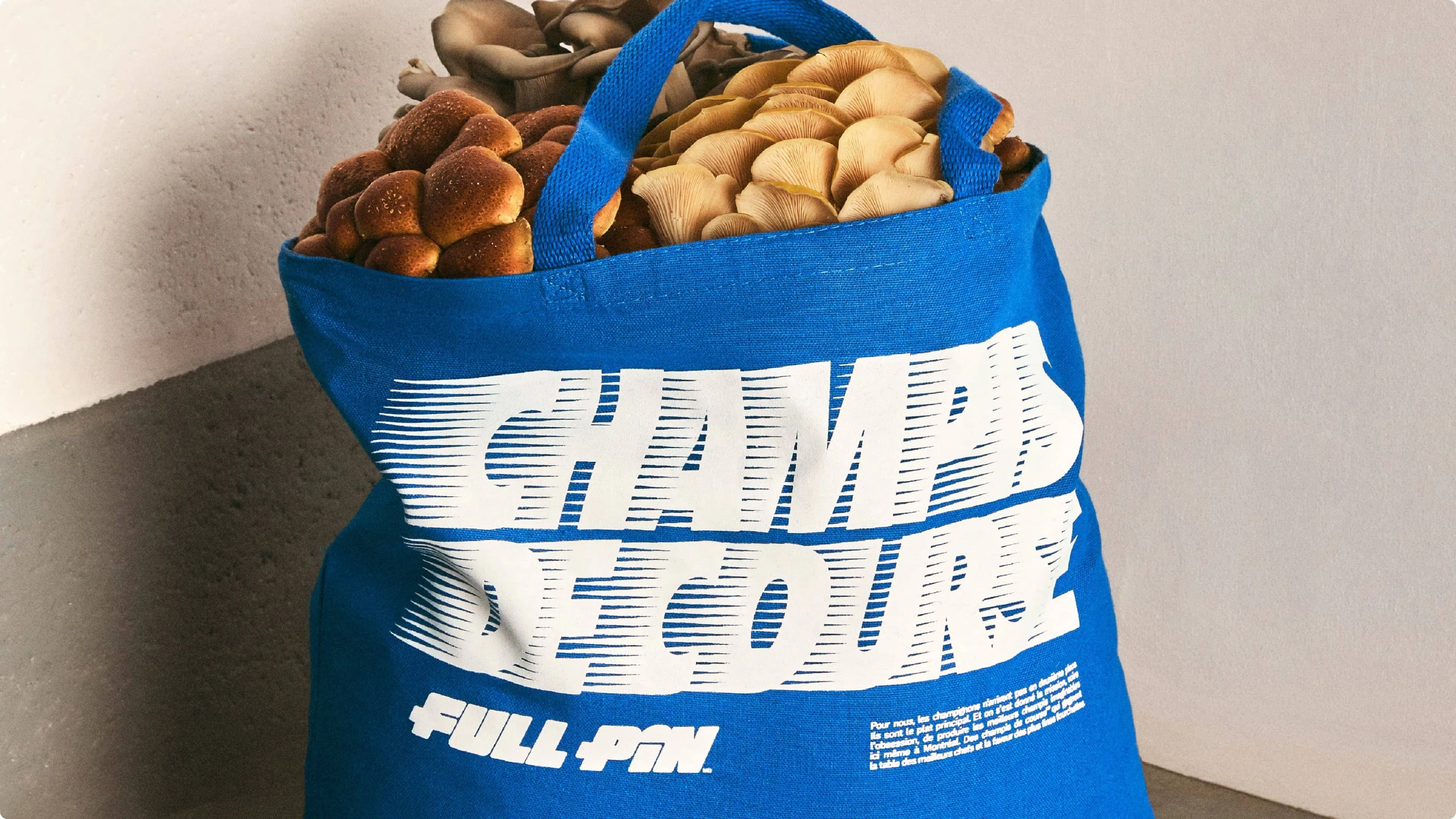 Blue tote bag filled with various mushrooms against a plain wall. The bag features bold white text, “CHAMPIS DE COURSE.”