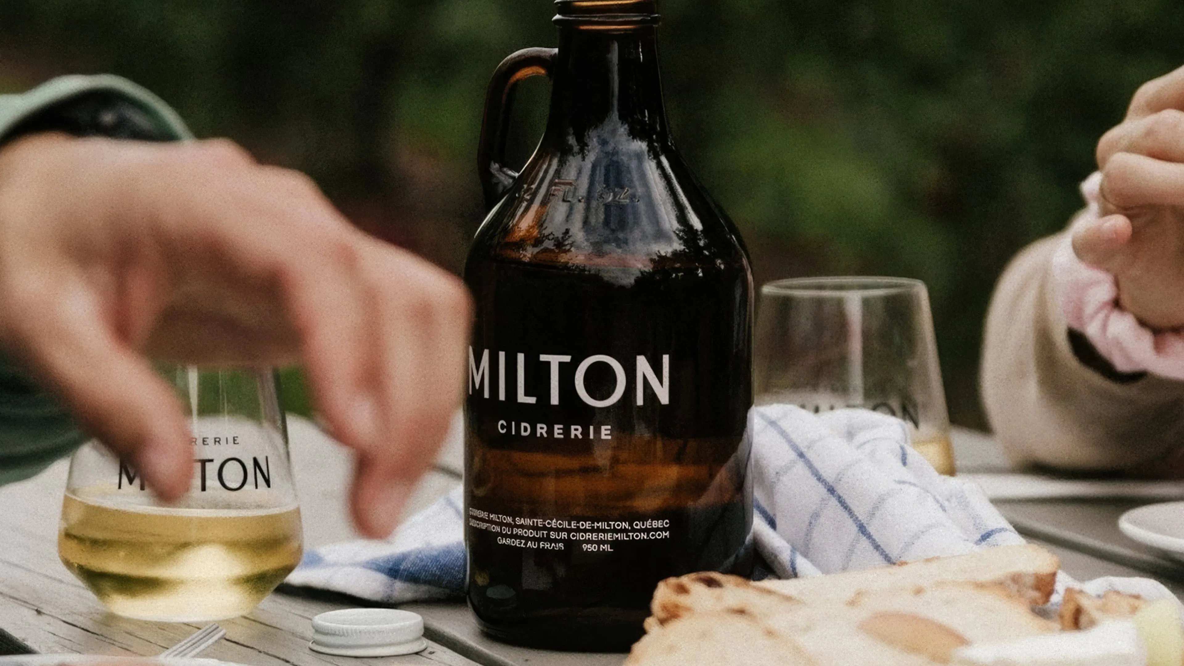 A brown bottle labeled "Milton Ciderie” sits on a table with glasses of cider, hands reaching for bread, and a relaxed outdoor setting in the background.