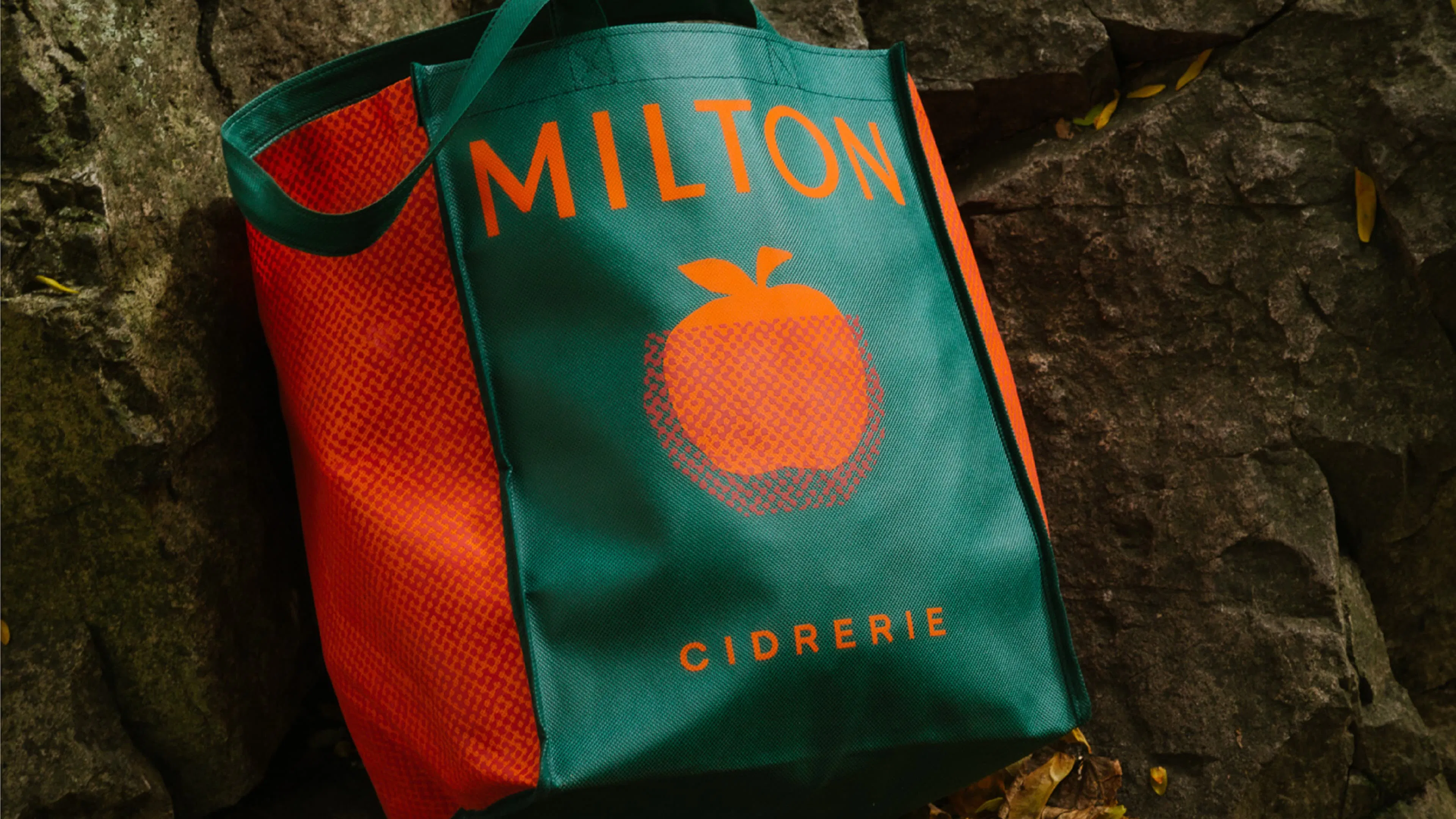 A green and orange MILTON CIDERIE tote bag rests against a rocky surface. The tote features an orange apple graphic.
