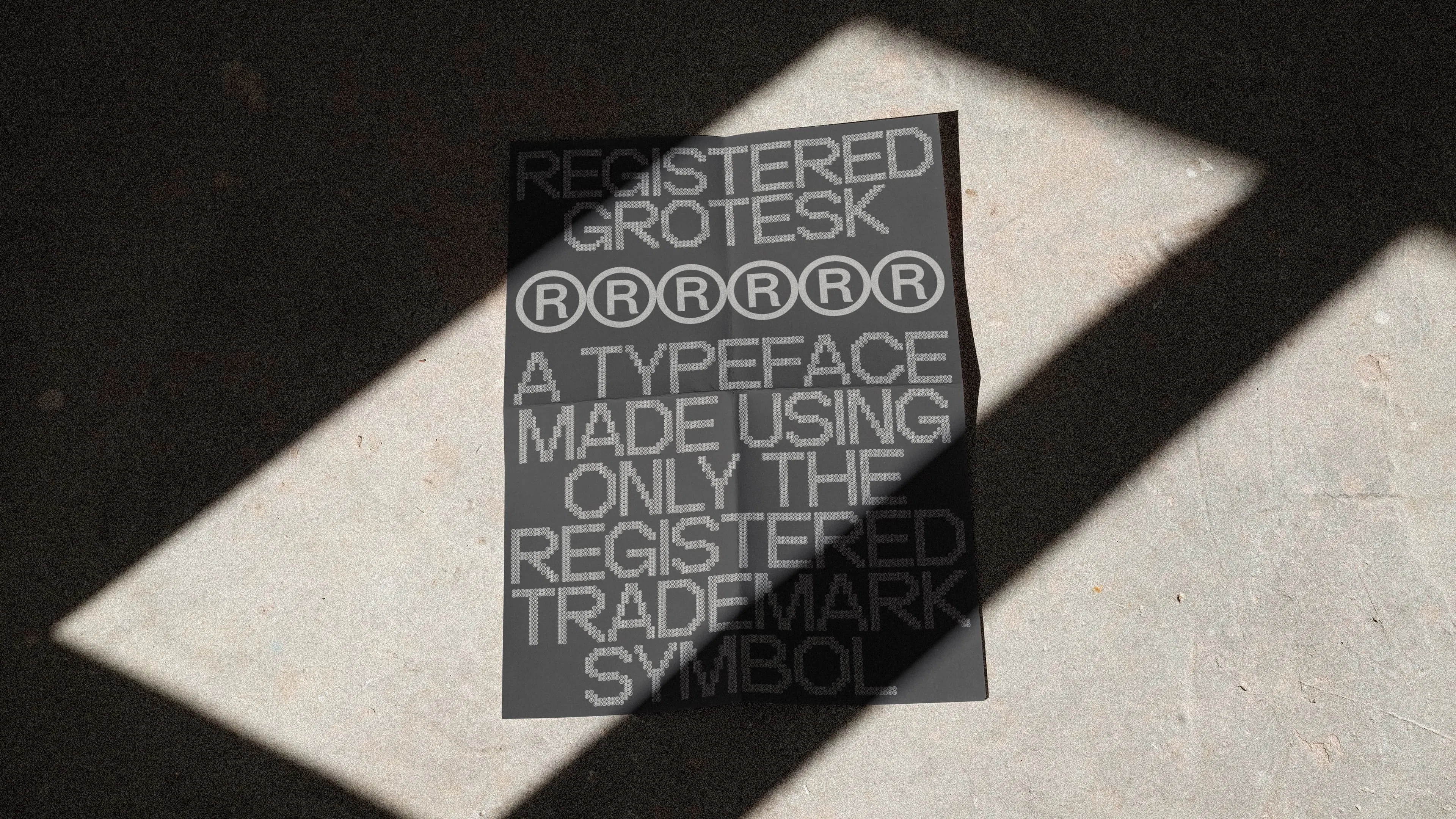 A black poster lies on a concrete floor, featuring white text reading "REGISTERED GROTESK" composed entirely of registered trademark symbols. Sunlight casts diagonal shadows across the poster, creating a dynamic contrast.
