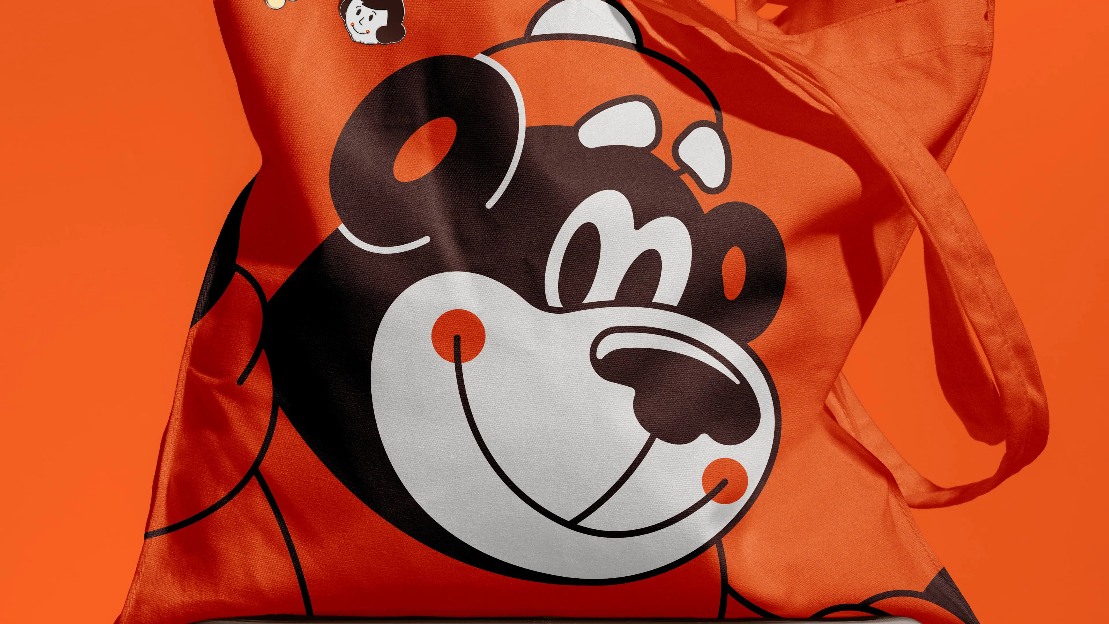Orange tote bag with a smiling cartoon bear face in black and white, resting on a dark stool. The background is a matching vibrant orange.