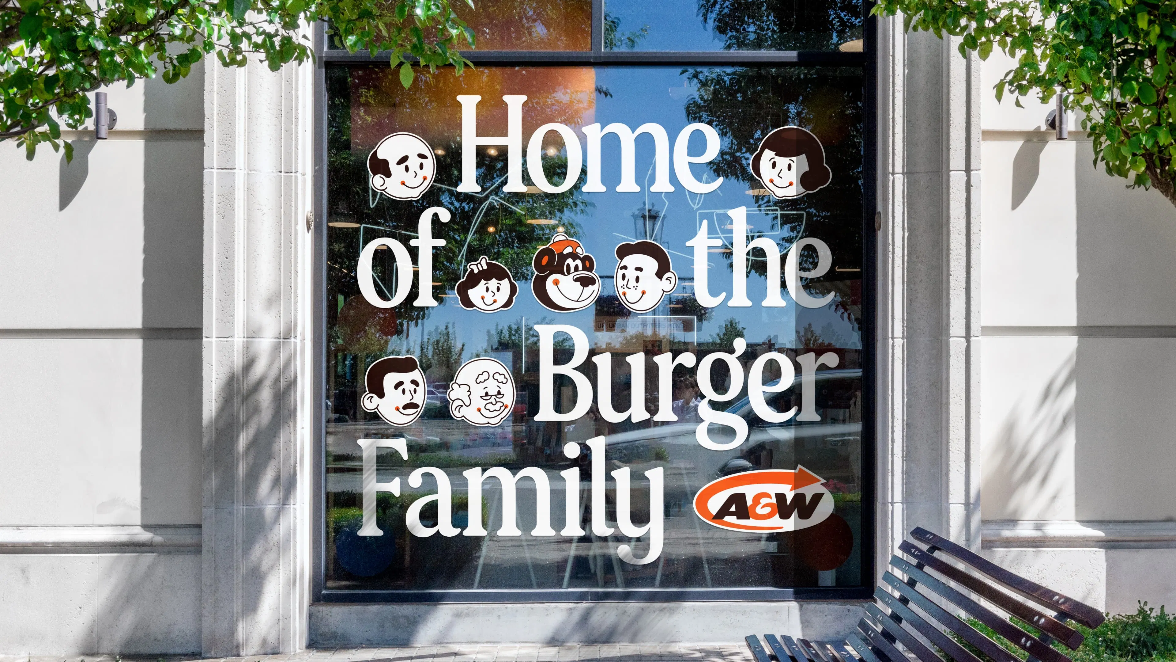 A restaurant window displays the phrase 'Home of the Burger Family' in bold white letters, surrounded by cartoon faces. Below, the A&W logo is visible. A wooden bench and tree branches frame the window.