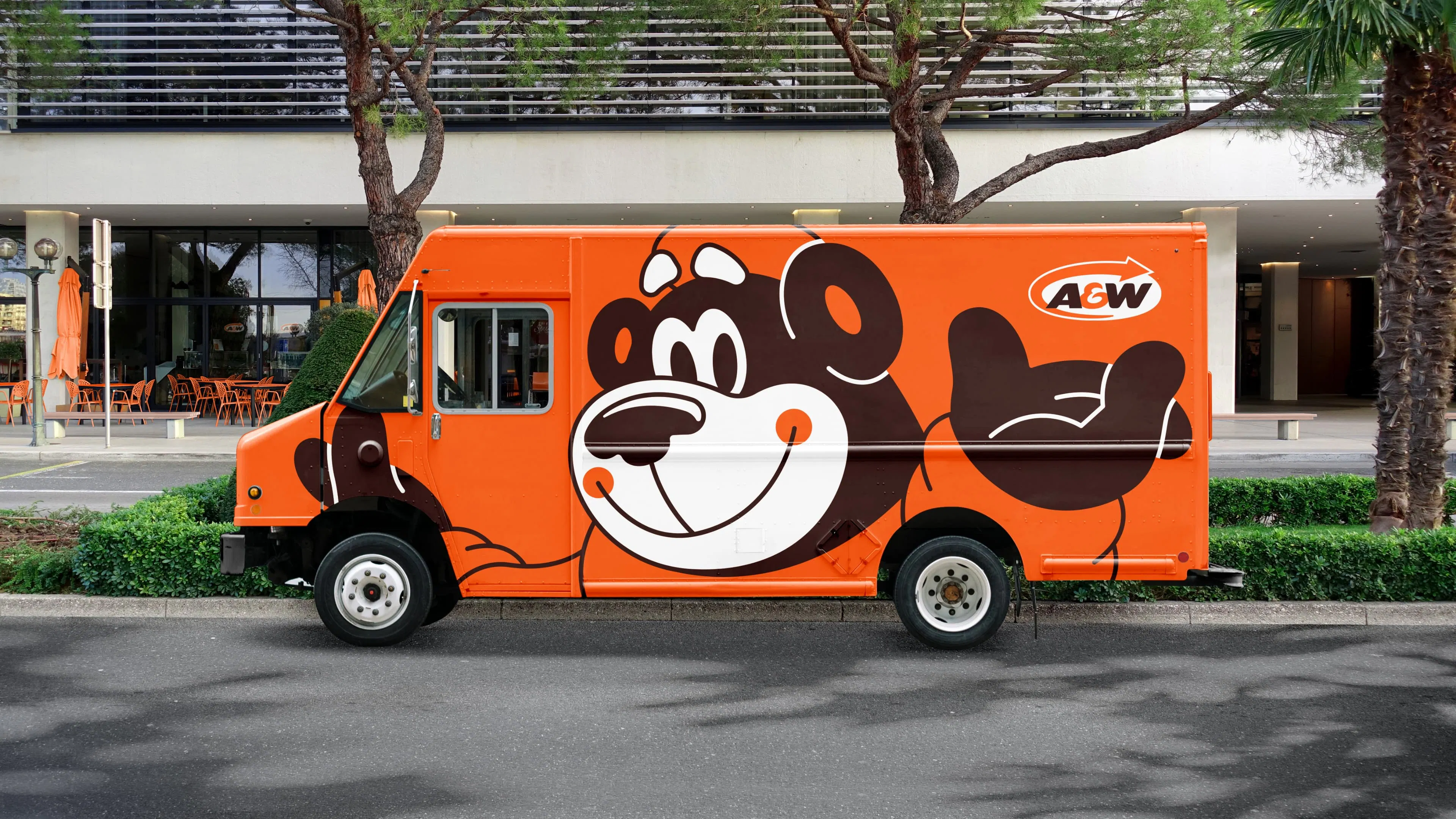 Orange food truck with a cheerful cartoon bear graphic, parked by a building with trees and outdoor seating.