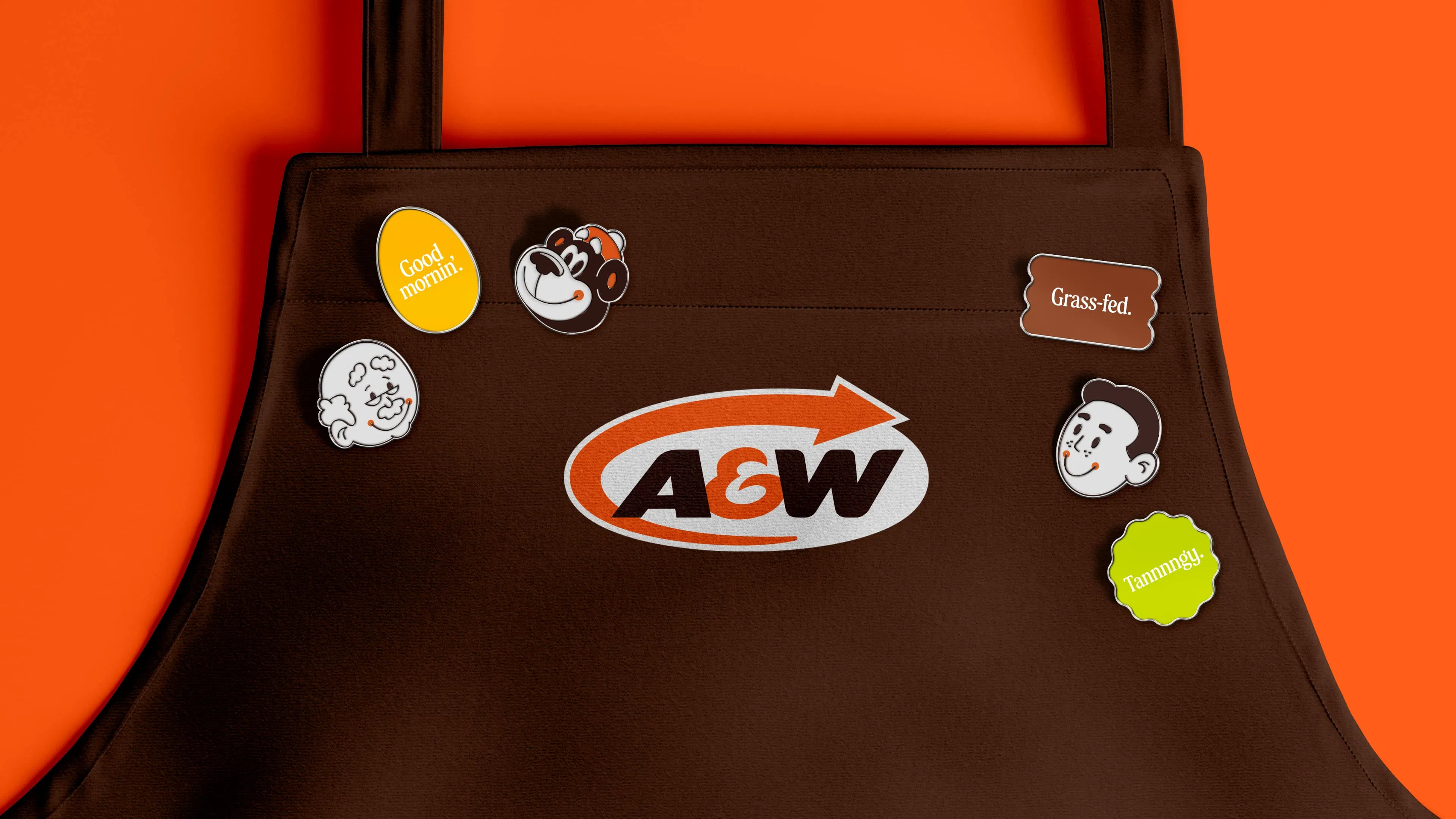 Brown apron with A&W logo in the center, adorned with playful pins: a bear head, "Good Mornin’", "Grass-fed" and smiling faces, against a bright orange background.