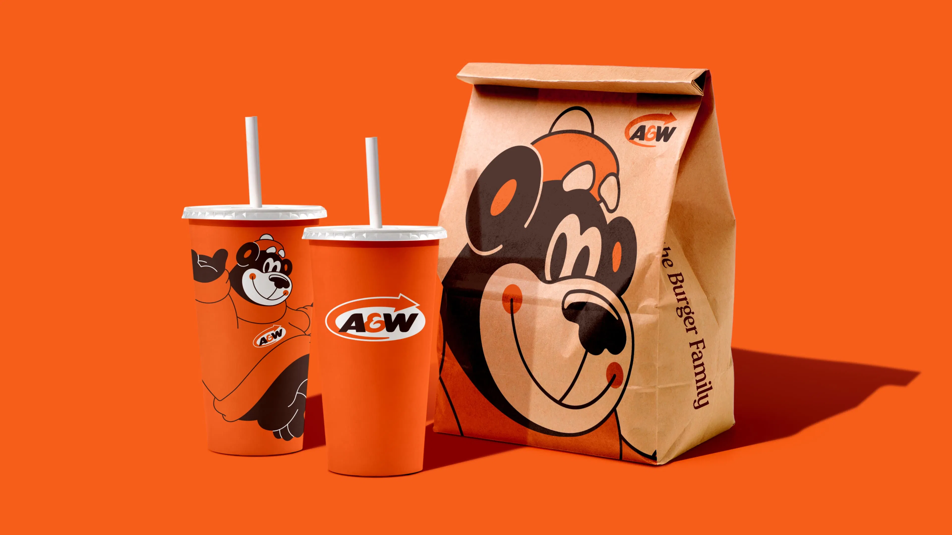 A&W fast-food packaging on an orange background. Two drink cups and a paper bag feature a smiling bear in a red hat.