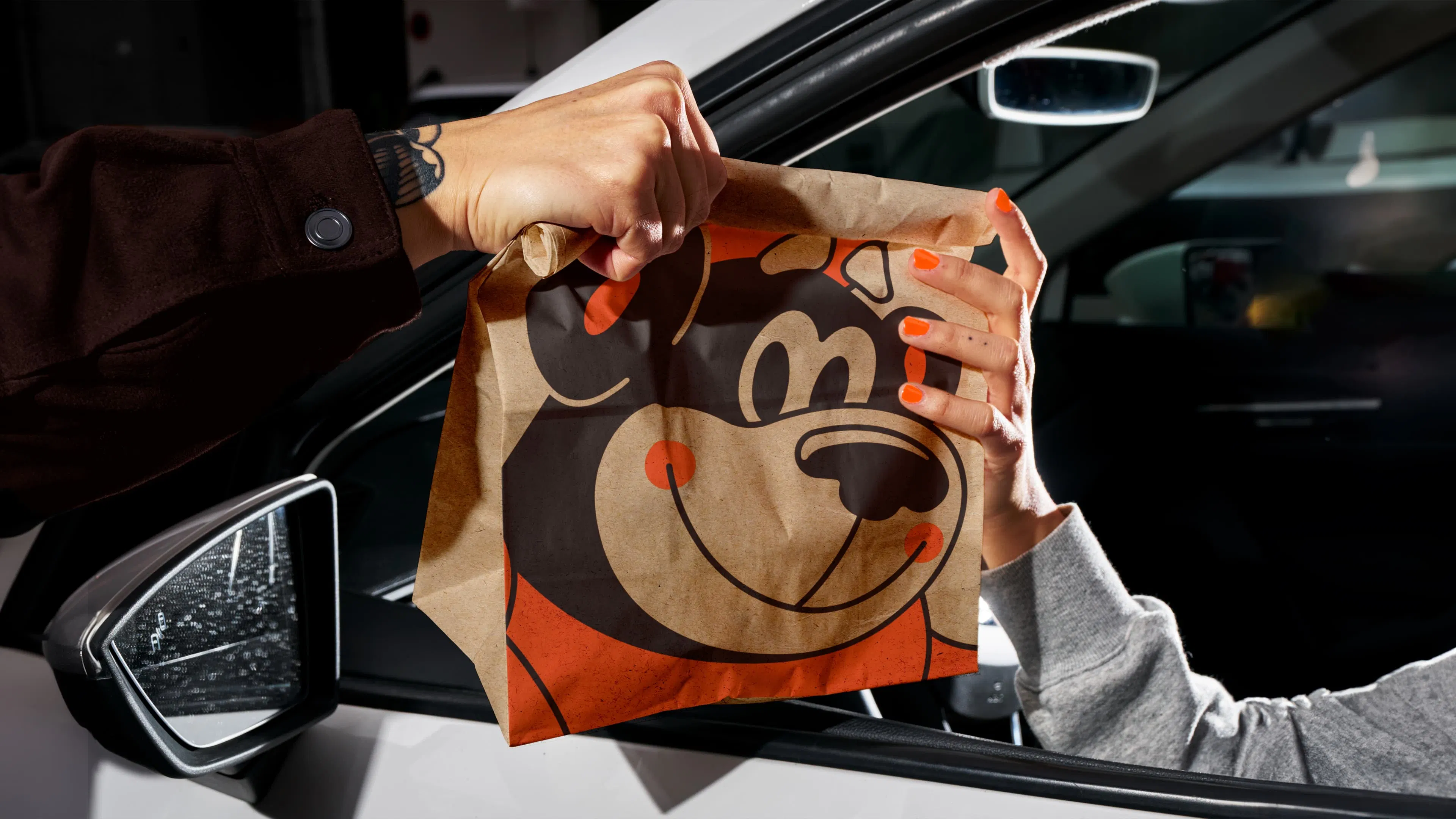 A hand with orange nails reaches out of a car window to receive a brown paper bag featuring a smiling bear logo.