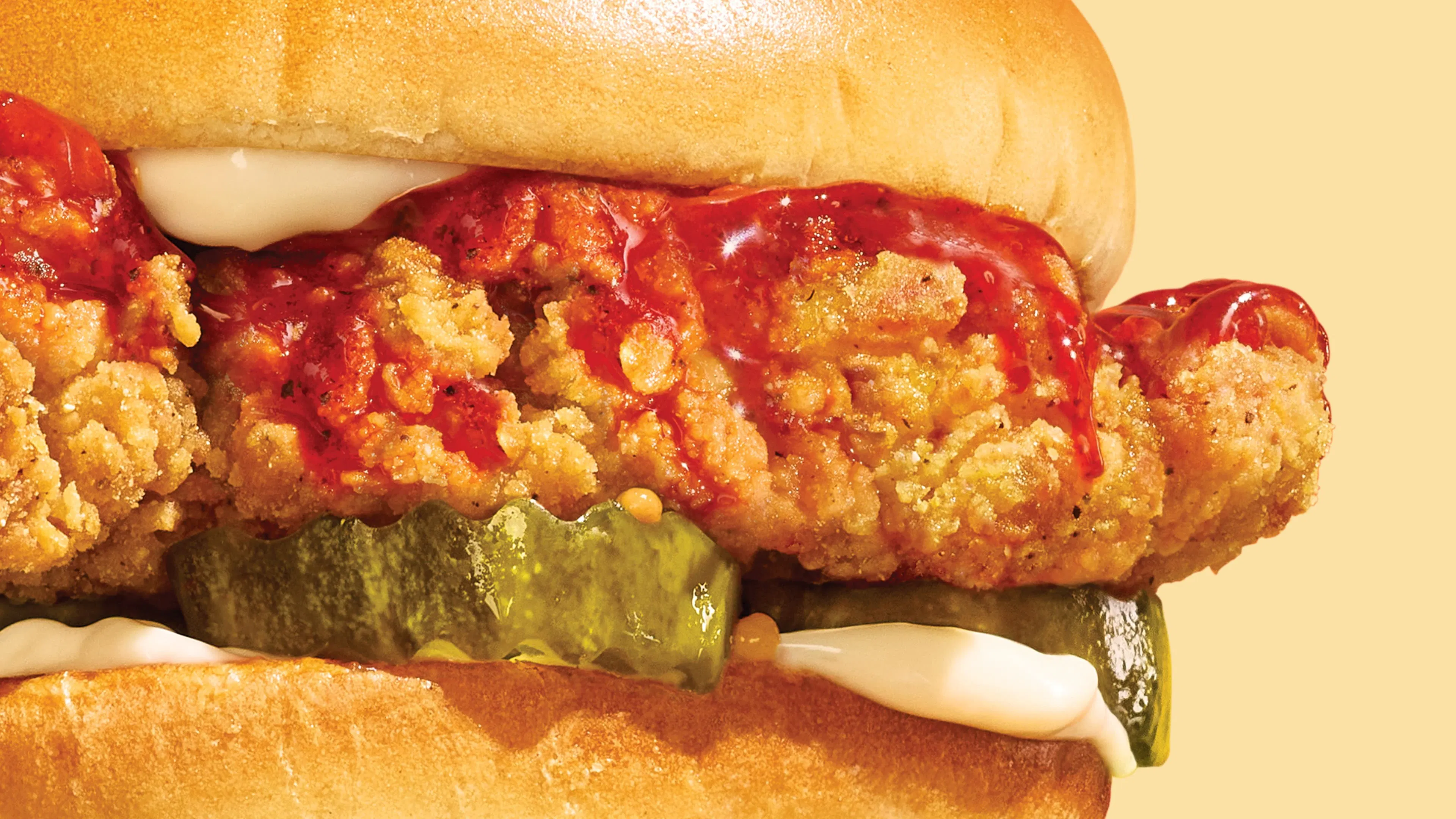 Close-up of an A&W crispy chicken sandwich with spicy sauce, pickles, and mayonnaise on a beige background.