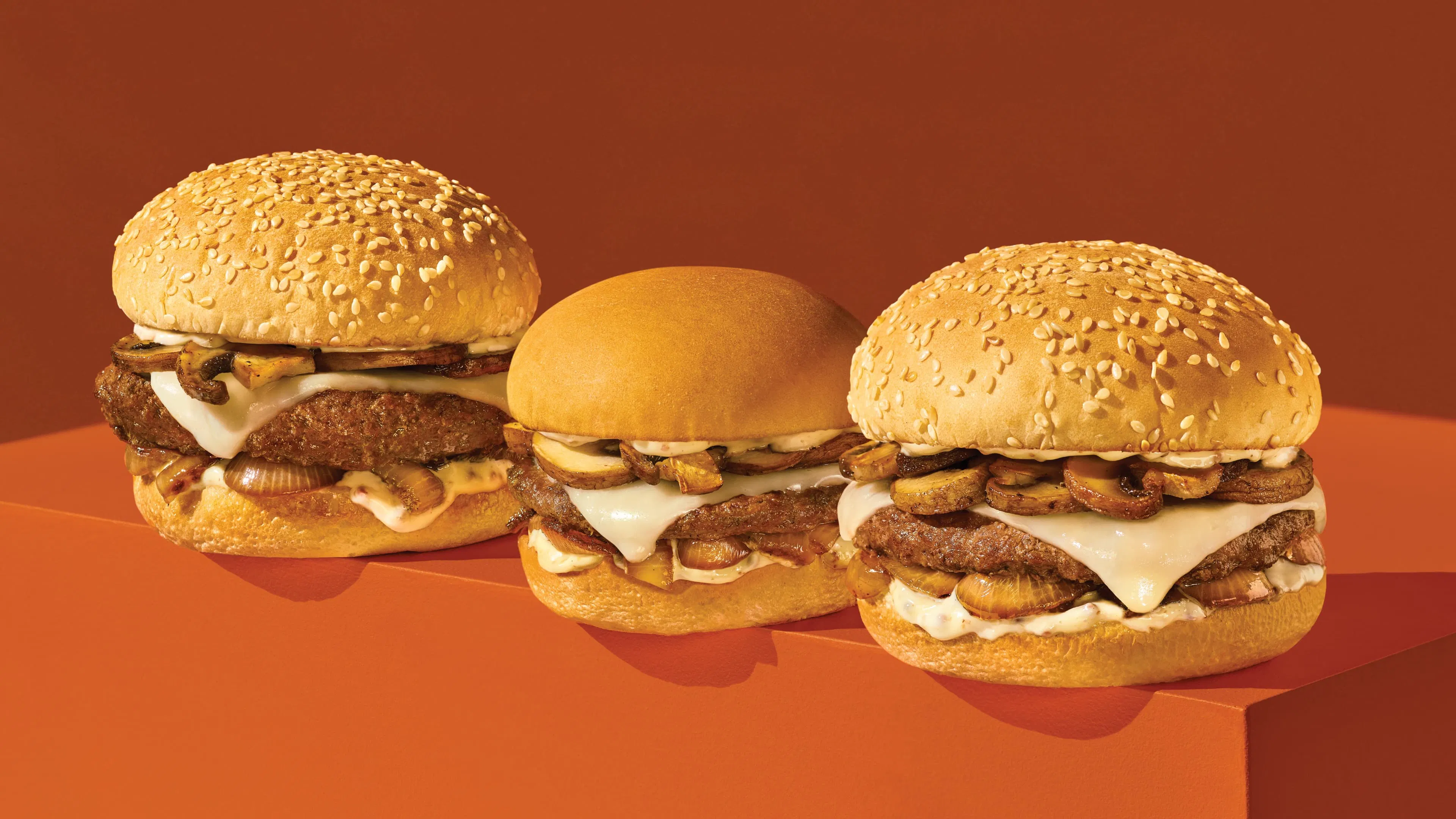 Three hamburgers on an orange surface, with toppings including melted cheese and mushrooms.