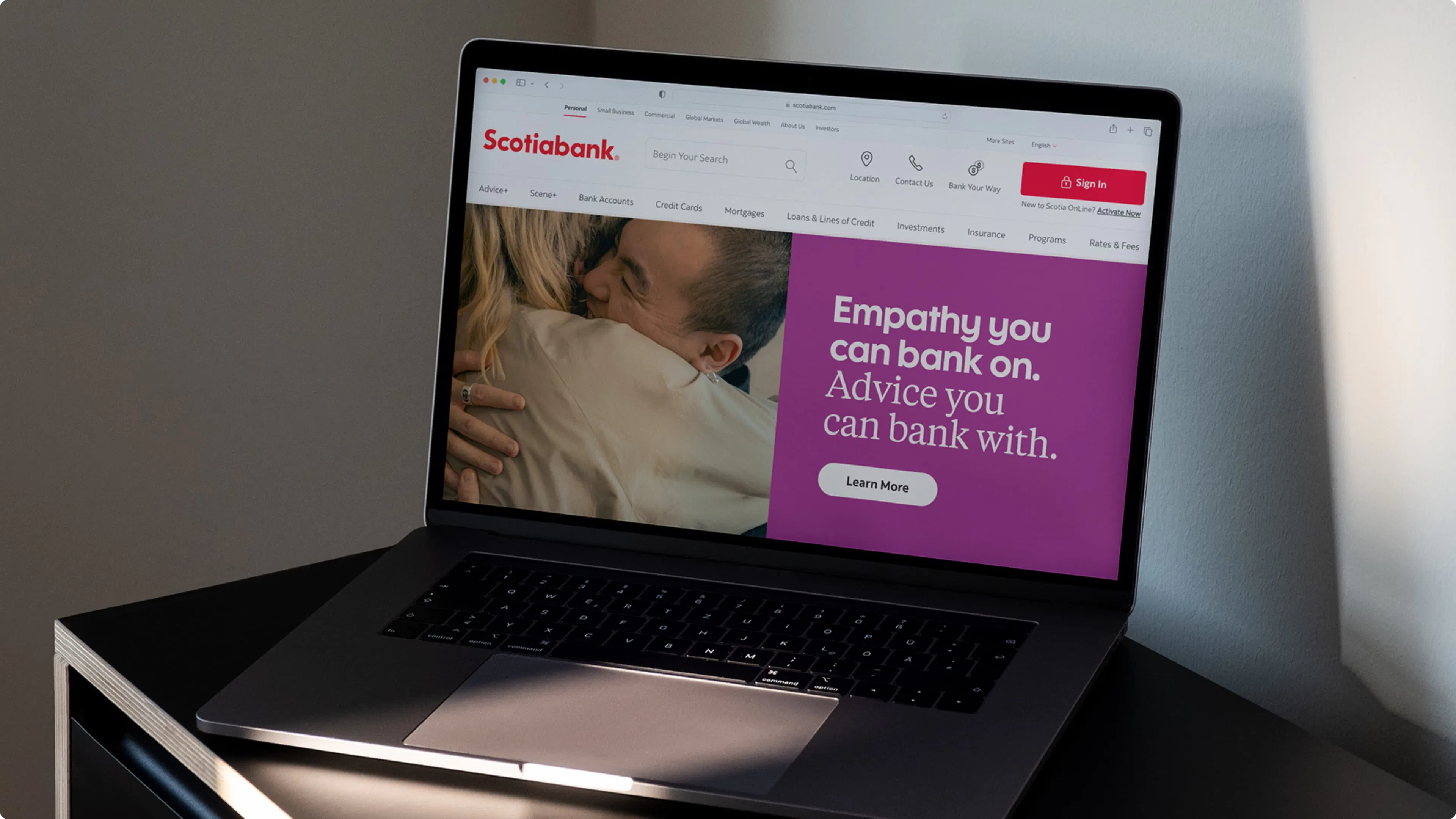 A laptop screen displaying a Scotiabank website with a purple banner stating, "Empathy you can bank on. Advice you can bank with." A human couple embraces on the left.