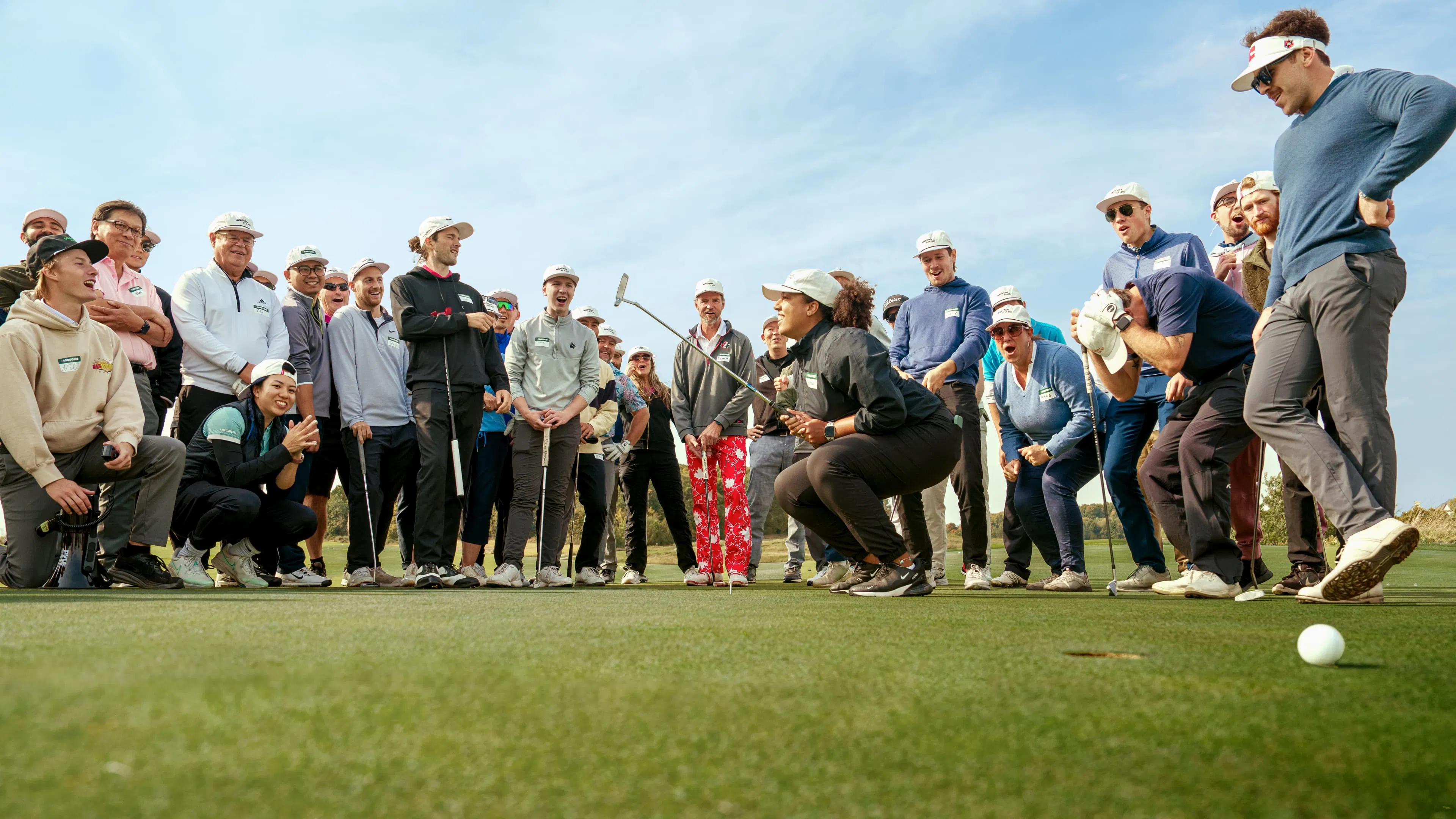 A large group of golfers, wearing hats, gather around a putting green, smiling and laughing. Two are poised as if performing a playful golf shot. The scene is lively and fun.