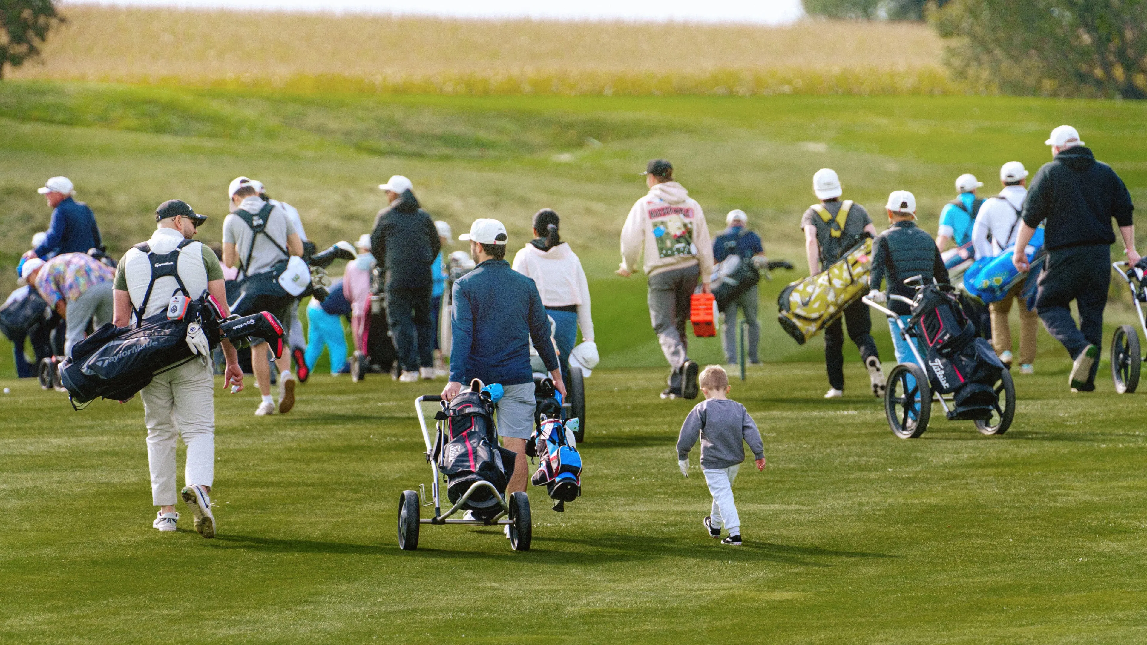 A group of golfers with bags and pushcarts walk along a lush green fairway. A small child in the foreground follows, adding a playful tone.