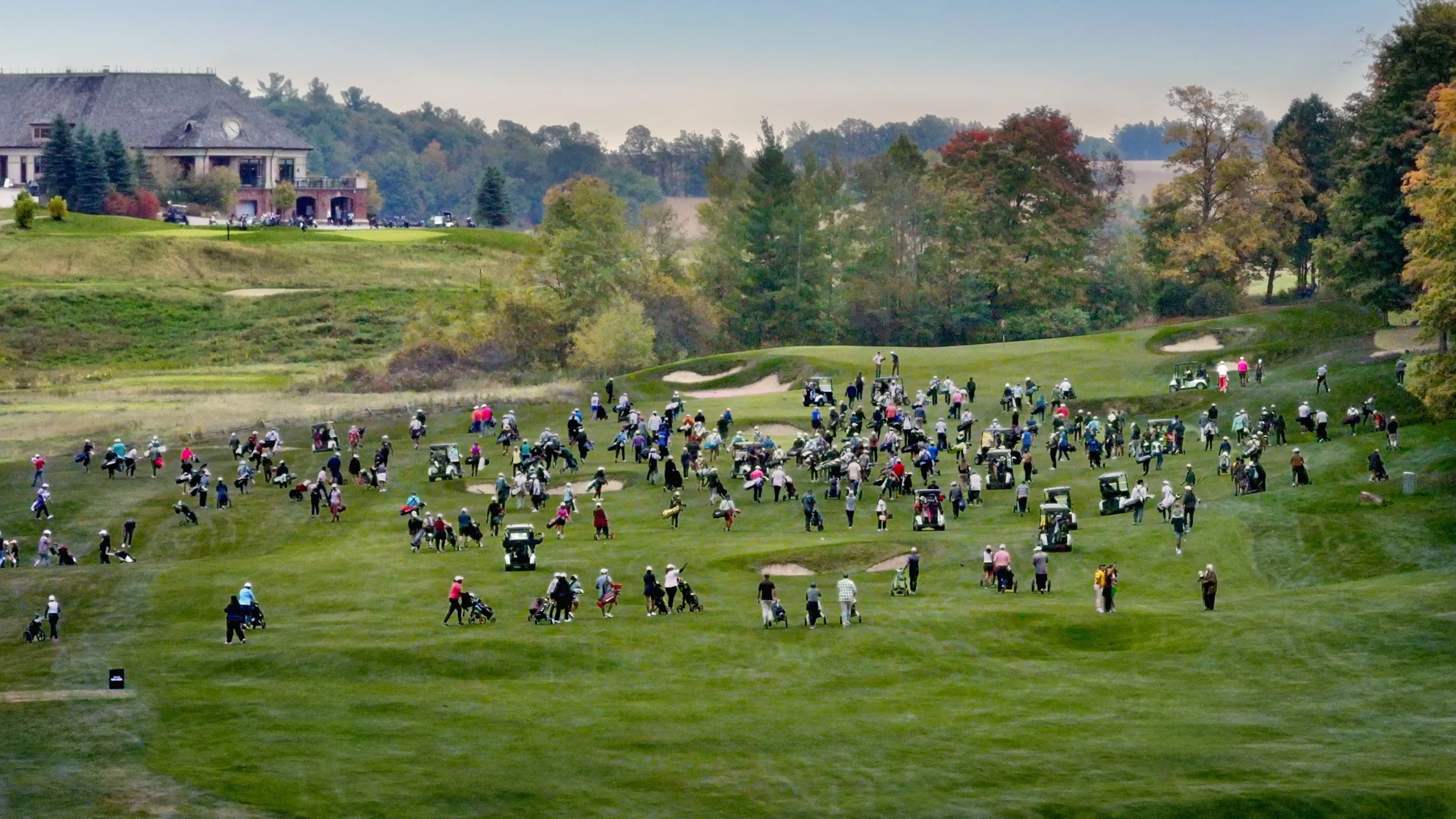Crowded golf course with numerous people dispersed across vibrant green fairways, set against a backdrop of autumn trees and an elegant clubhouse.
