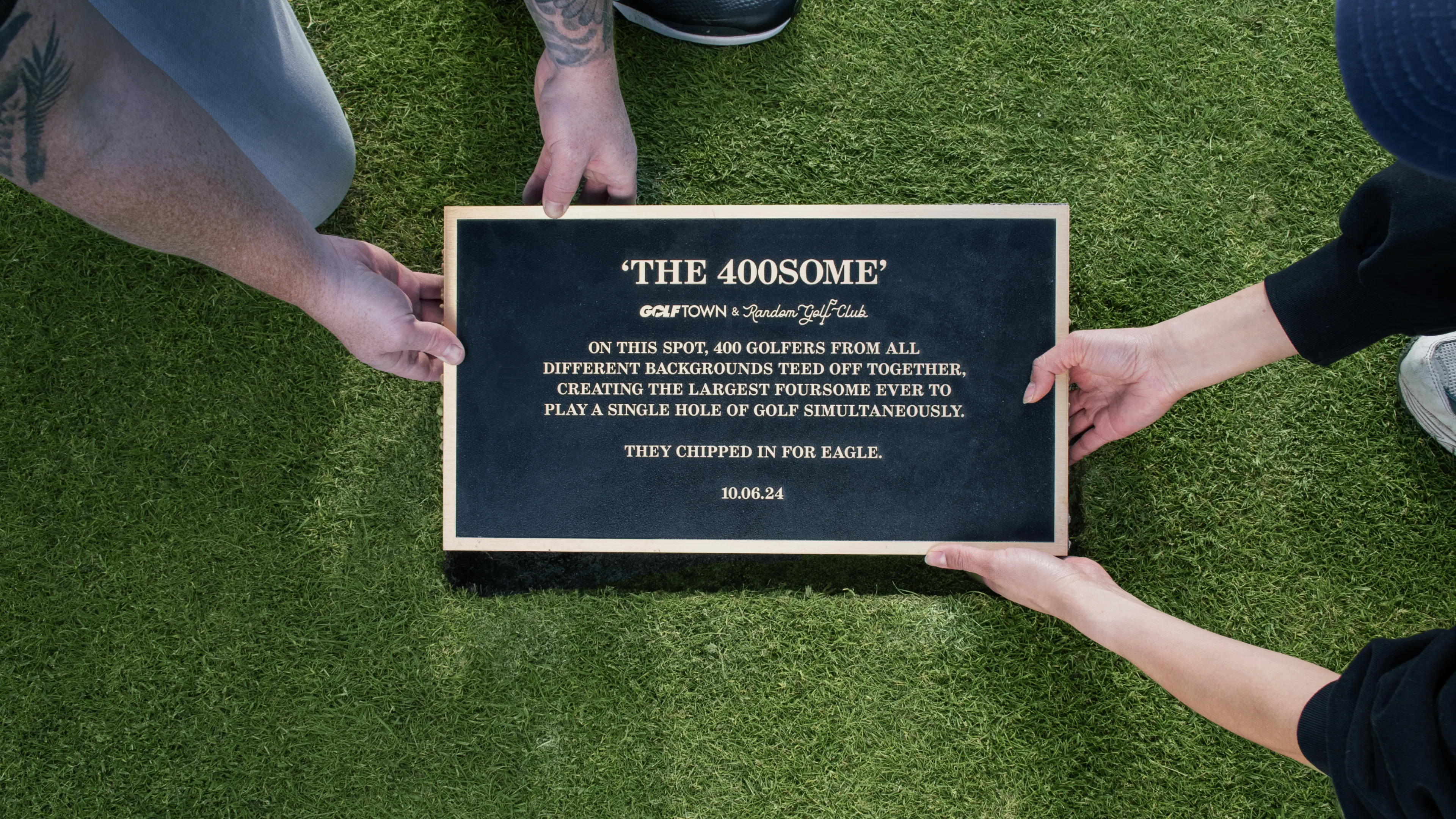 Two individuals place a plaque on grass, titled "The 400some." It commemorates 400 golfers playing together. The mood is celebratory and historic.