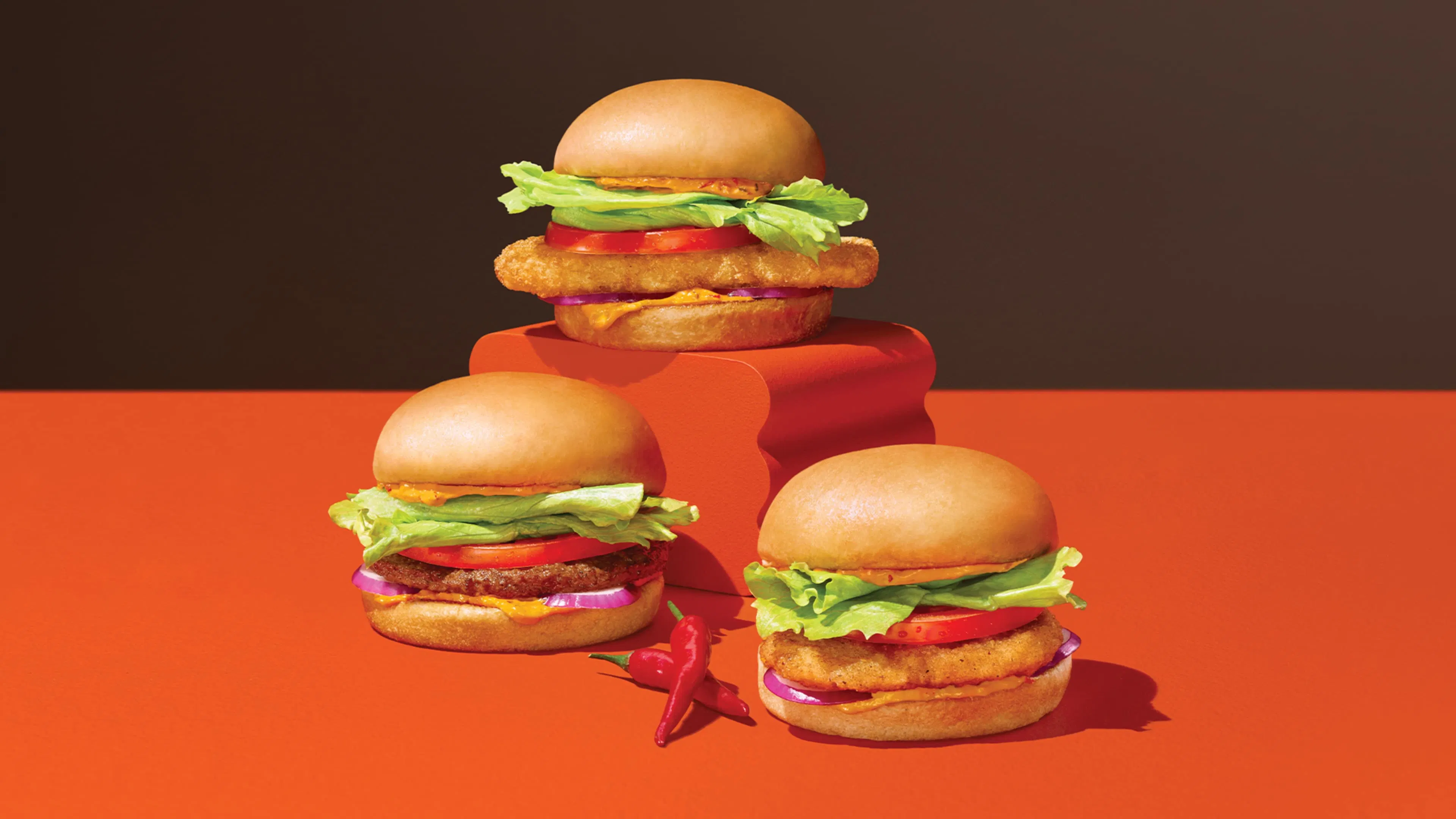 Three A&W Spicy Piri-Piri burgers are arranged on an orange surface, with one elevated in the center. Each burger contains lettuce, tomato, and crispy fillings, one each with seasoned breaded chicken breast, a beef patty and crispy hash brown potato patty. Two red chillis are placed in front.