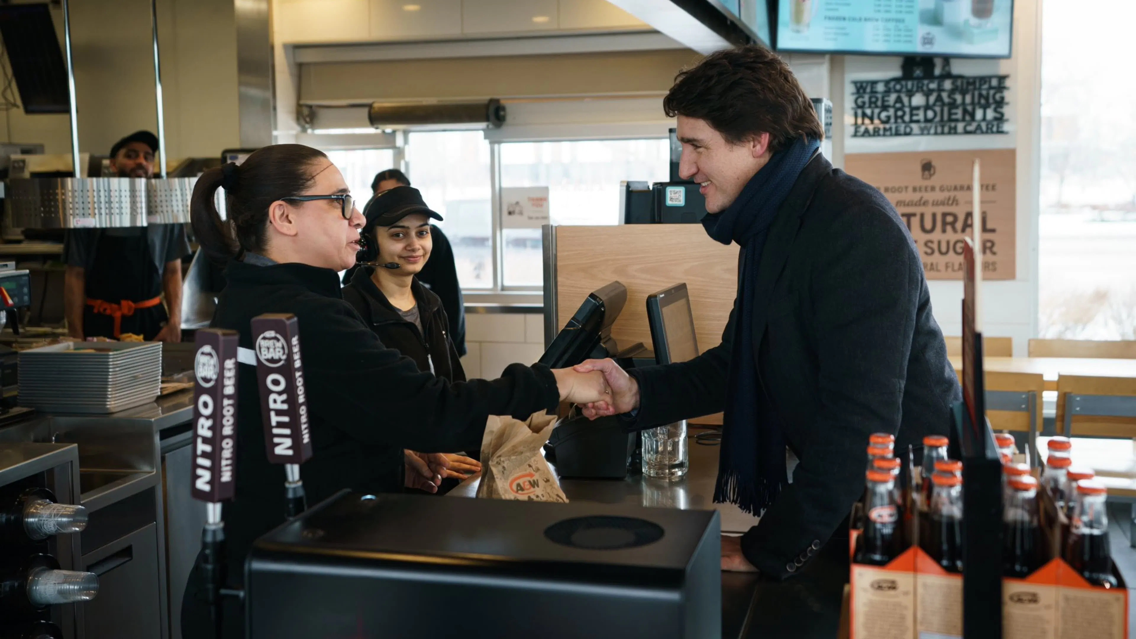 Former Prime Minister Justin Trudeau of Canada wears a dark coat and shakes hands with a female A&W franchise operator across the counter, smiling. The scene is warm and welcoming.