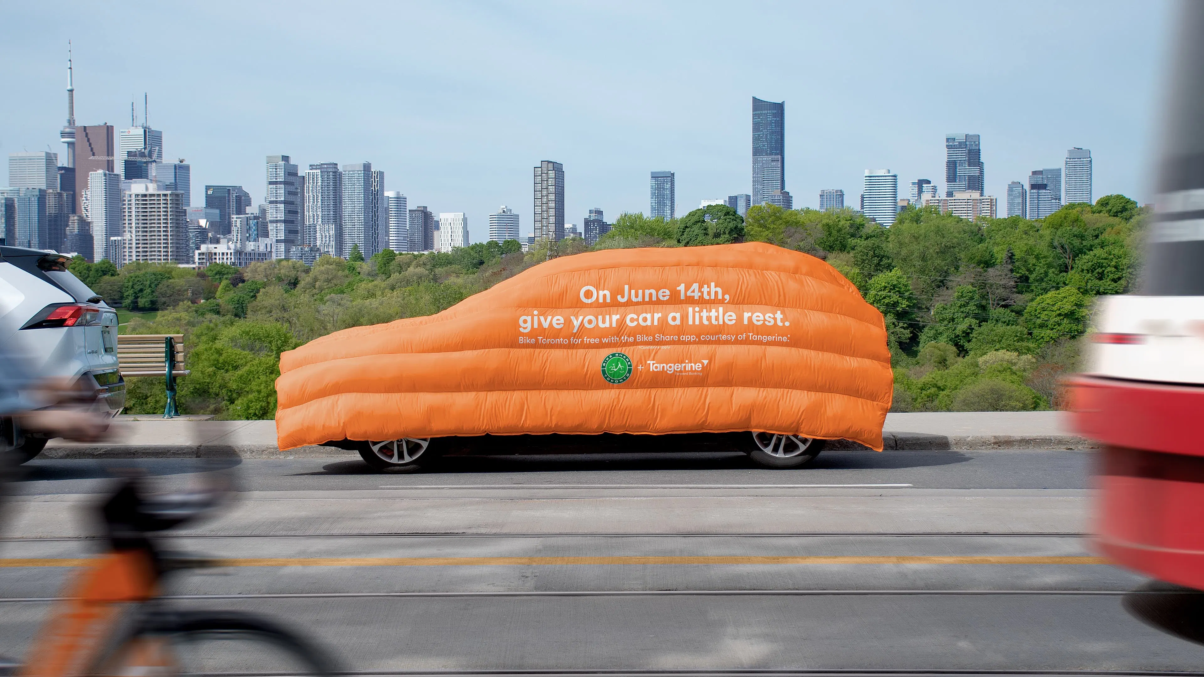 A parked car on the side of the street is covered with a comically large orange duvet bearing the text, "On June 14th, give your car a little rest." A cyclist on a Bike Share Toronto bike passes in the foreground with moving traffic. The Toronto skyline is behind the car.