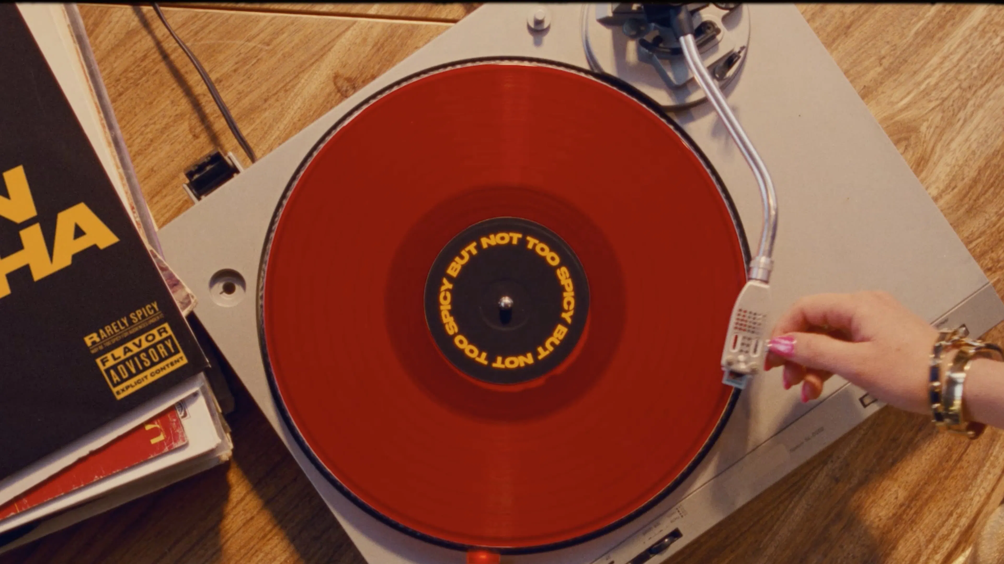A hand adjusts the tonearm on a spinning red vinyl record player, set against a wooden table with stacked album covers nearby, evoking a retro vibe. The vinyl reads “Spicy But Not Too Spicy”