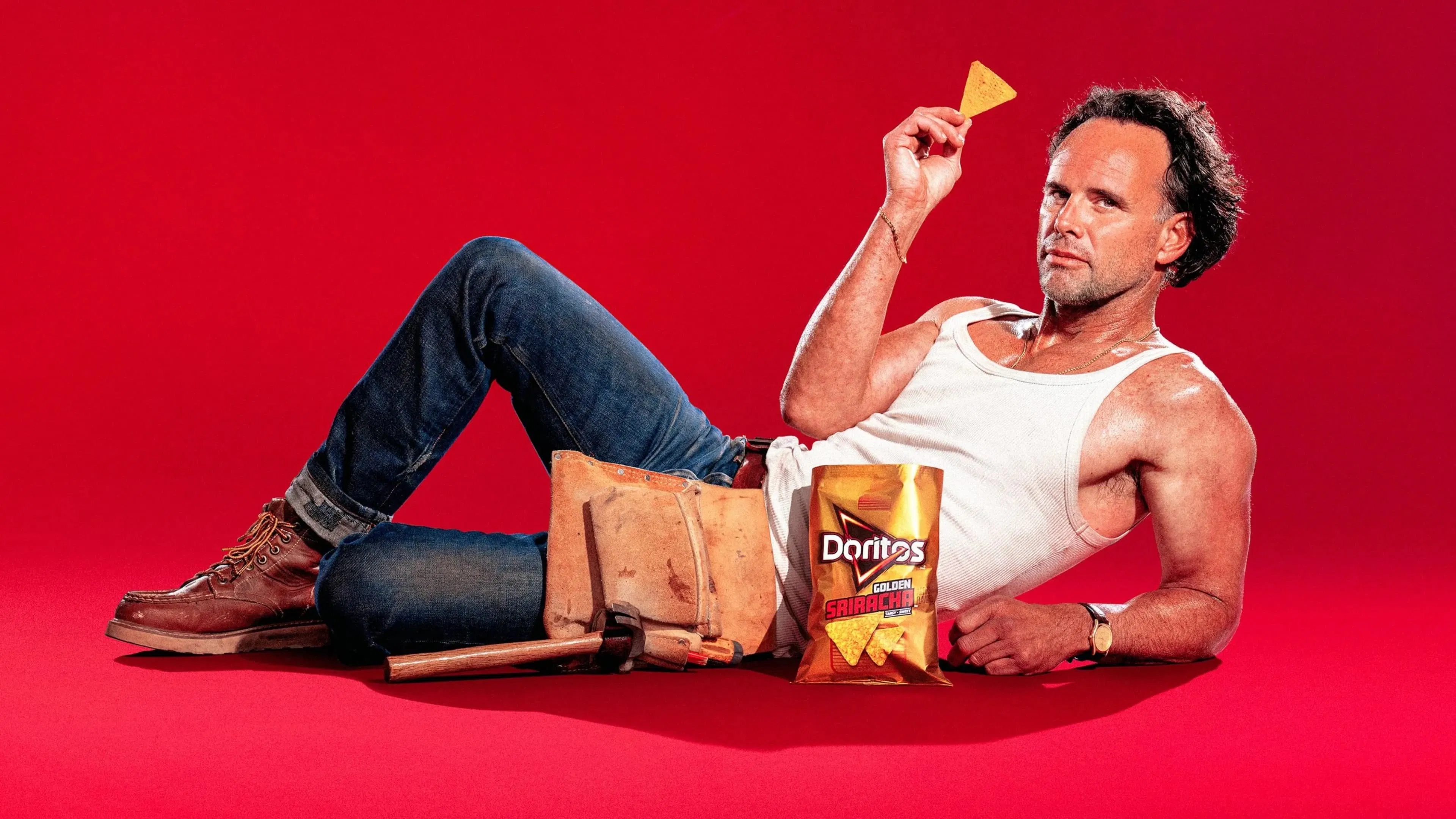 Actor Walter Goggins is in character as a plumber, wearing a white tank top and jeans, a tool belt and work boots. He is casually reclining on a red background, holding a Doritos chip, with a bag of Doritos Golden Sriracha in front of him. His gaze is suggestive and flirty in nature.