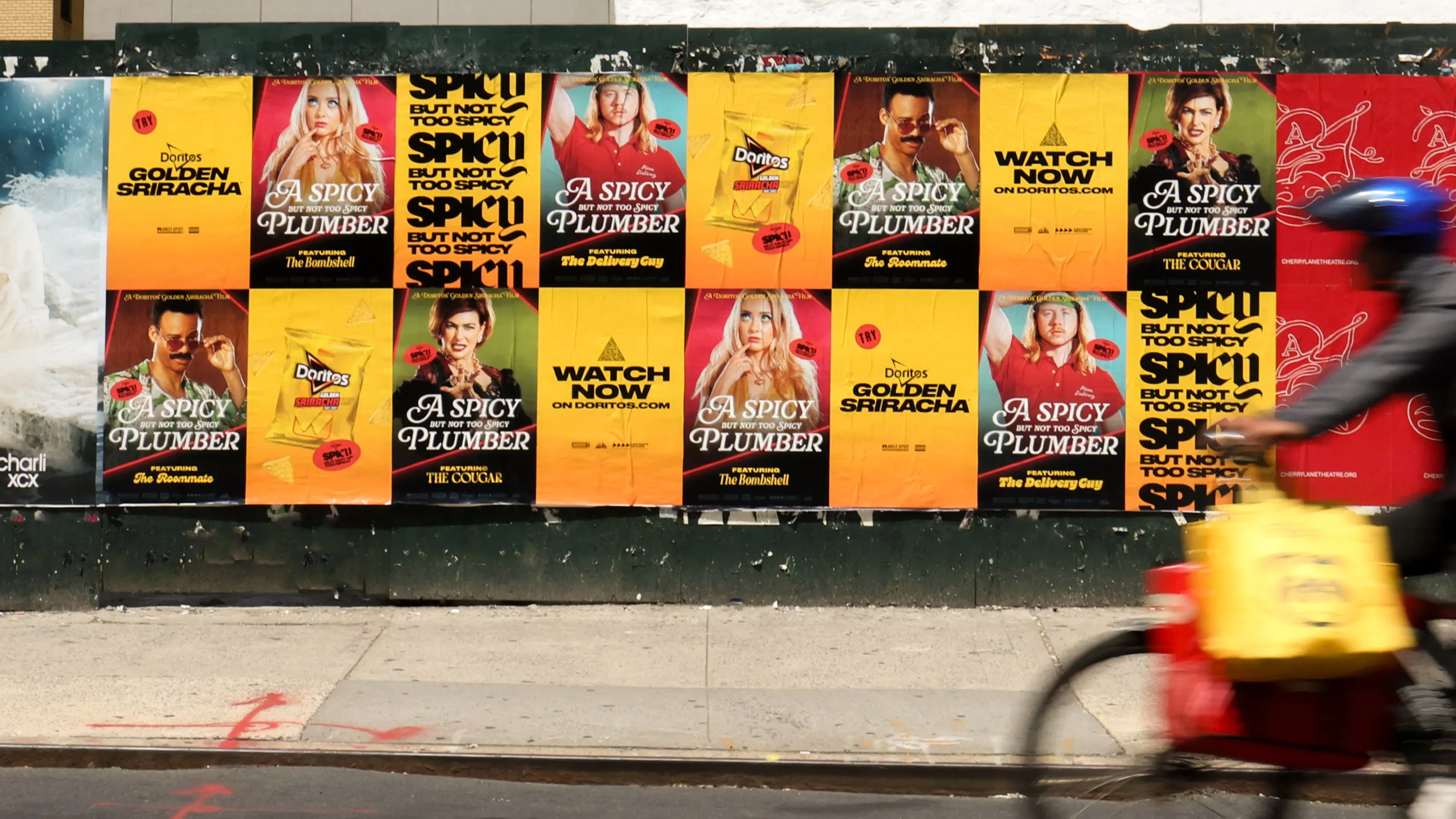 A poster wall of ads displays out of home ads promoting the Doritos Golden Sriacha through a fake movie called, A Spicy Plumber, featuring different characters part ofthe commercial. A cyclist in motion passes by quickly, adding dynamic energy.