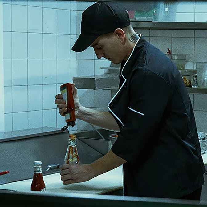 A kitchen staff member in a black uniform is seen putting non-Heinz ketchup into a Heinz ketchup bottle in a busy kitchen environment