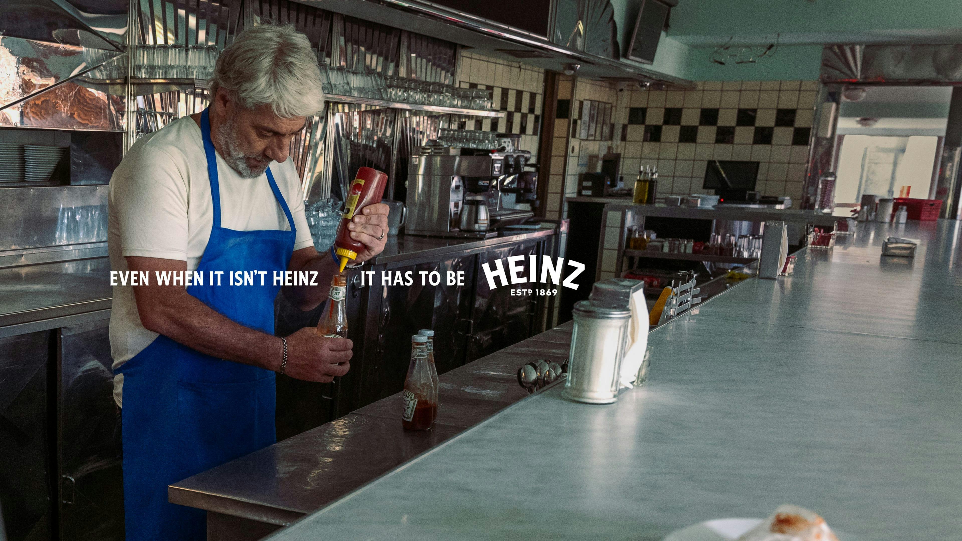 Un homme avec les cheveux blancs et un tablier bleu est en train de mettre du ketchup qui n’est pas du Heinz dans une bouteille de ketchup Heinz dans un restaurant de type casse-croûte.