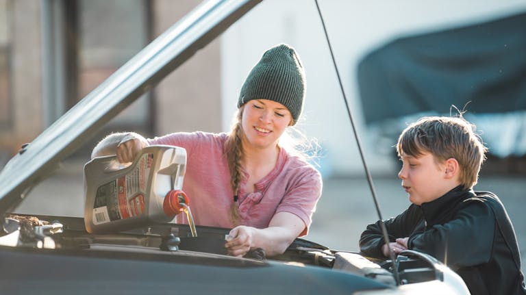 Eine Frau mit Mütze füllt Motoröl in den Motor eines Autos ein, während ein Junge daneben steht und zuschaut. Perfektes Beispiel für das Nachfüllen von Motoröl bei einem Ölwechsel.