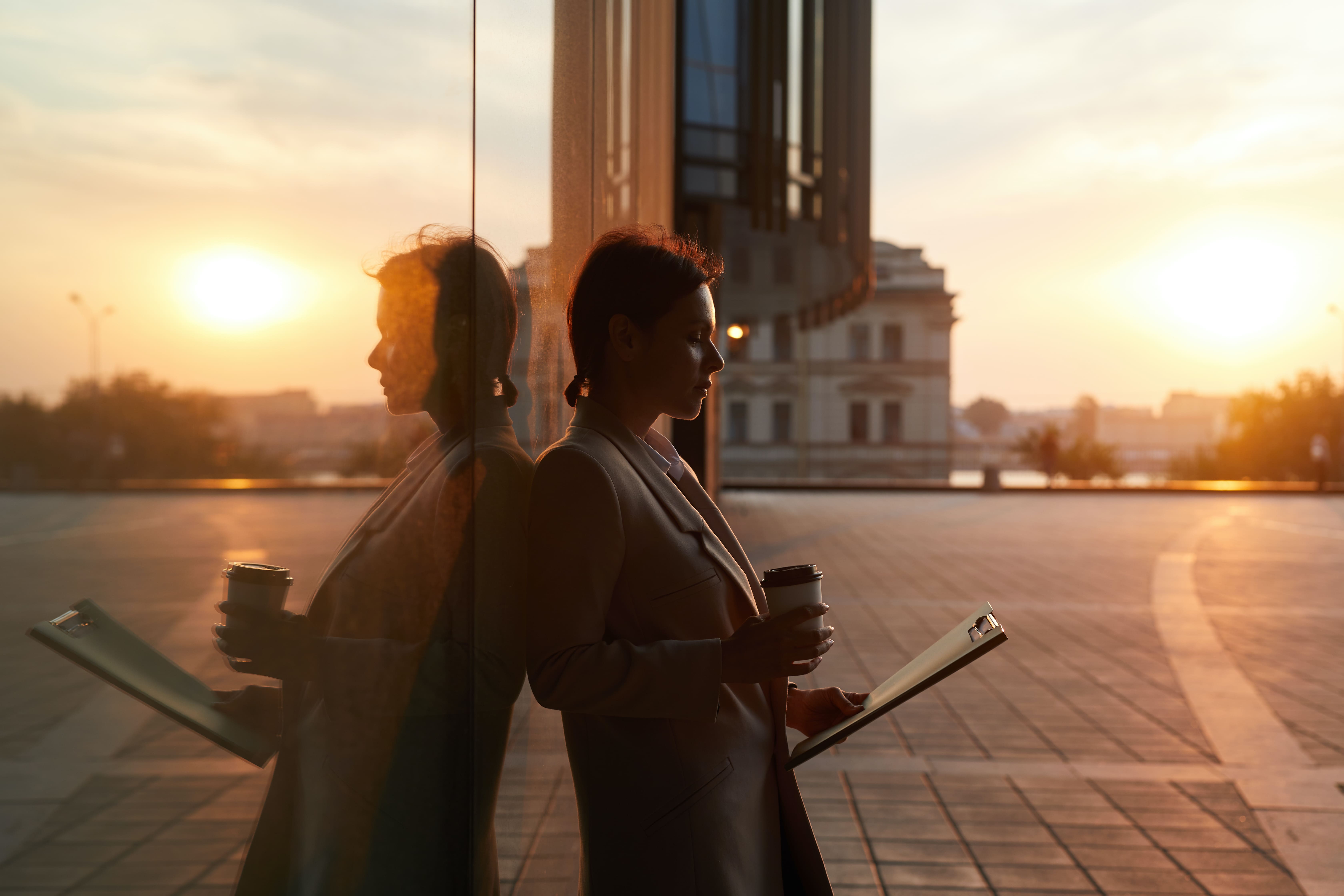 Frau lehnt auf einem Platz in der Abendsonne an einer Glasfassade.