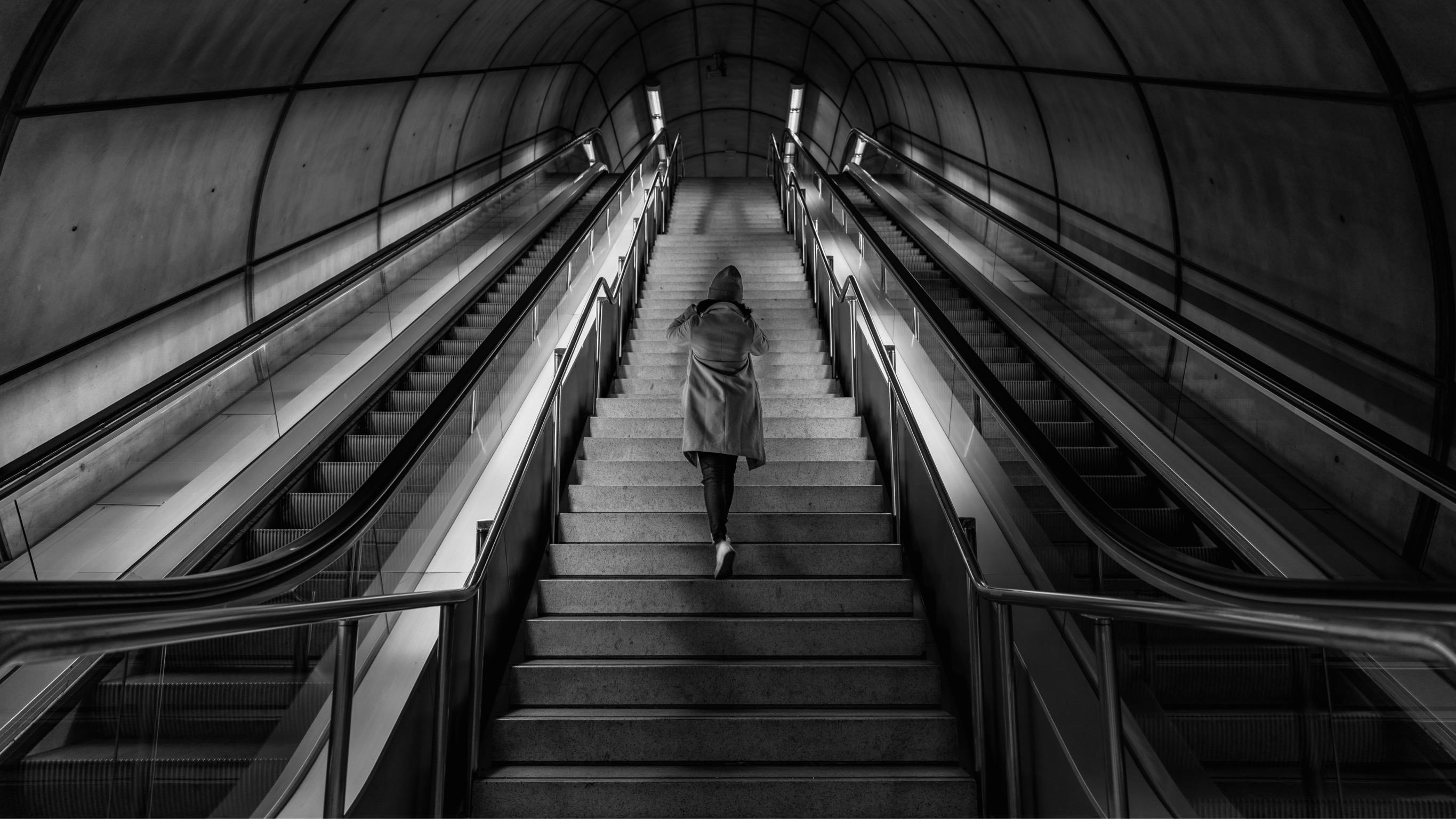 Eine Frau von hinten, die zwischen zwei Rolltreppen eine lange Treppe hochläuft.