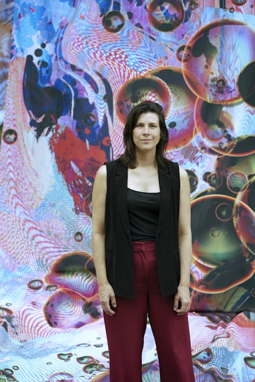 A portait of Kayva van Gangelen. A person stands in front of a colourful artwork depciting abstract bubbles.