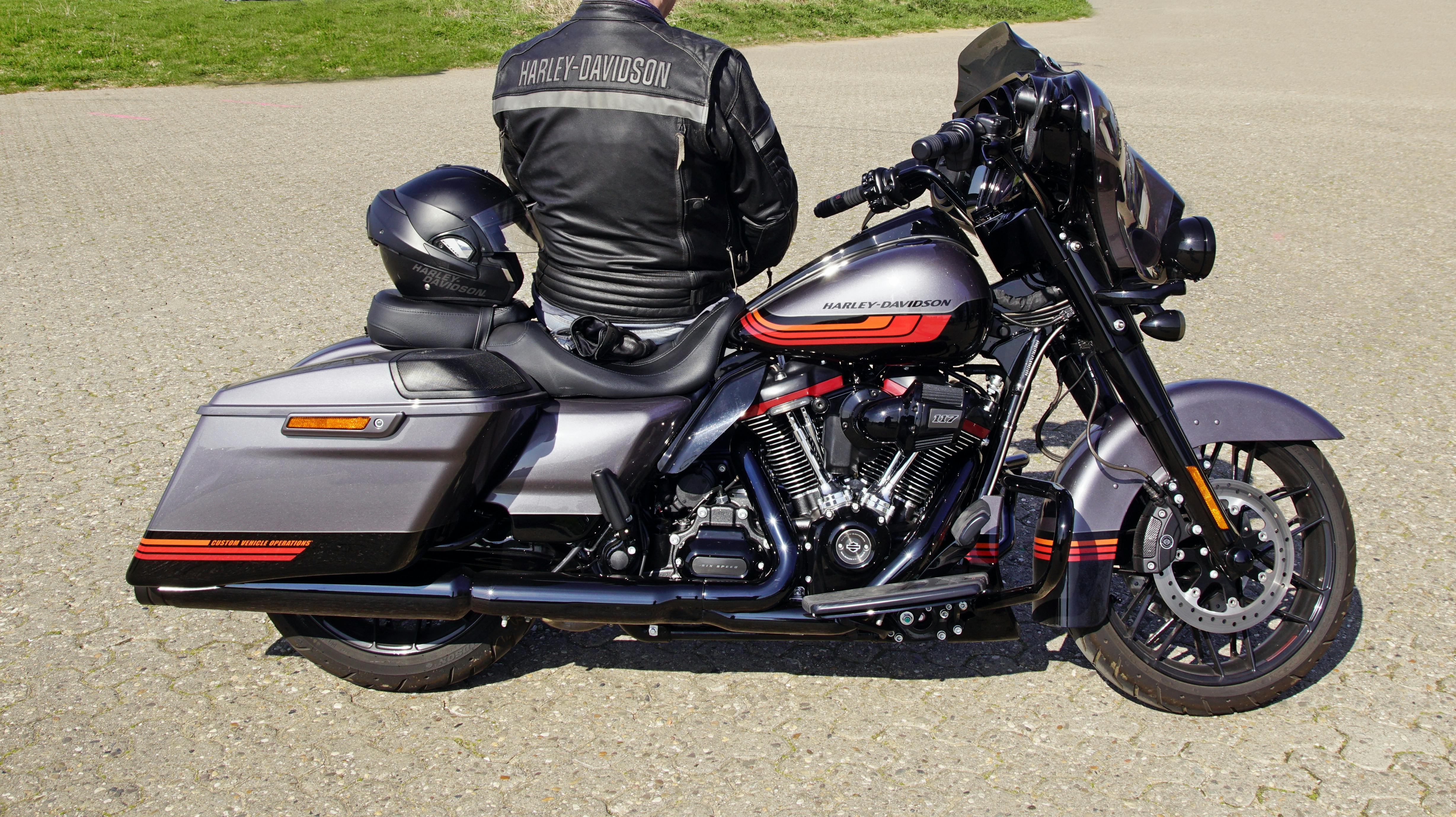 Man in a leather Harley jacket sitting on a Harley Davidson Road King motorcycle in a parking lot