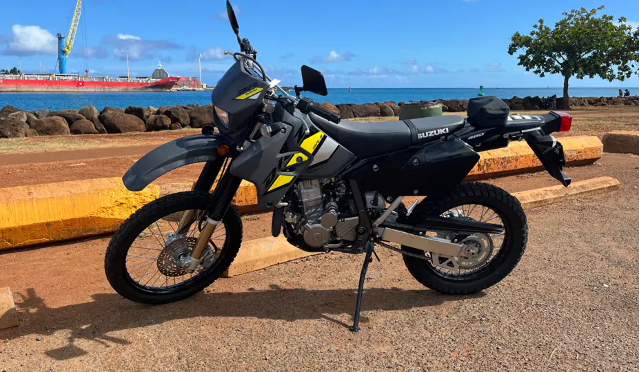 Foto de uma 2021 Suzuki DR-Z400s disponível para aluguel através do Riders em Hawaii, estacionada ao longo de uma costa com um grande navio ao fundo