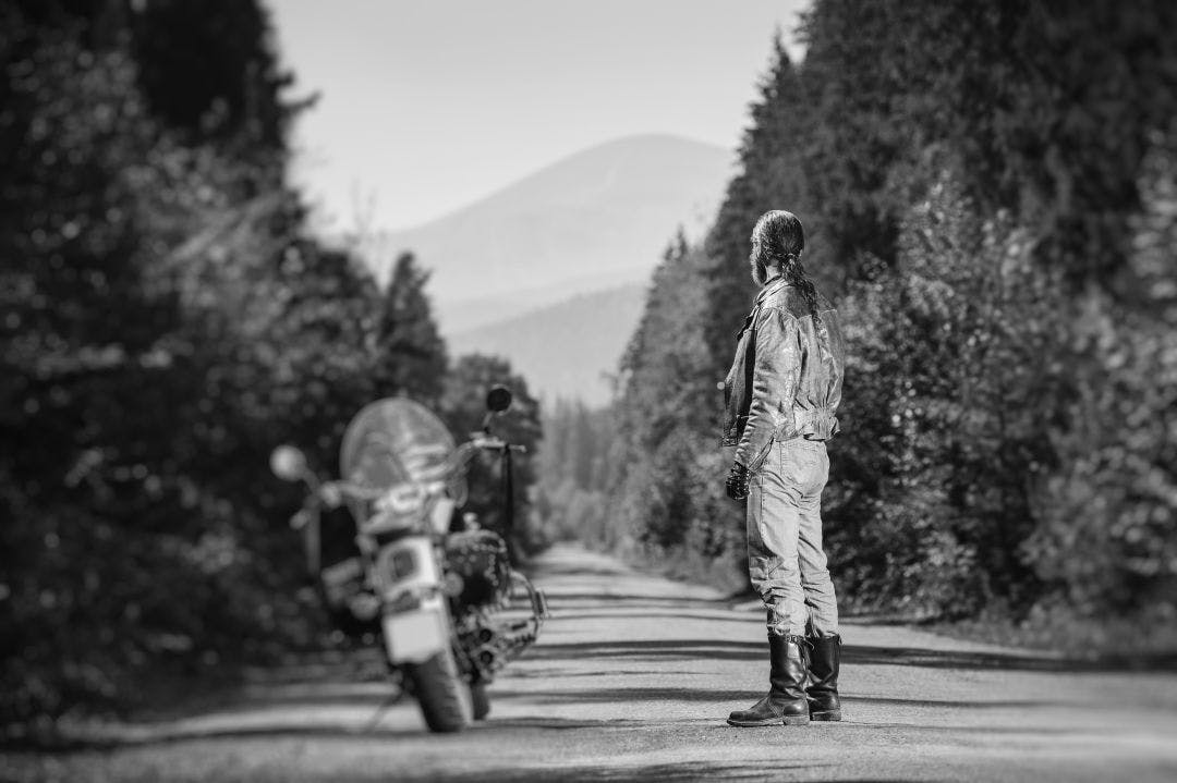 Biker with long hair wearing leather jacket jeans leather boots and gloves standing near his custom made cruiser motorcycle on the open road. Looking into distance Guide to International Motorcycle Riding & Rentals in the USA
