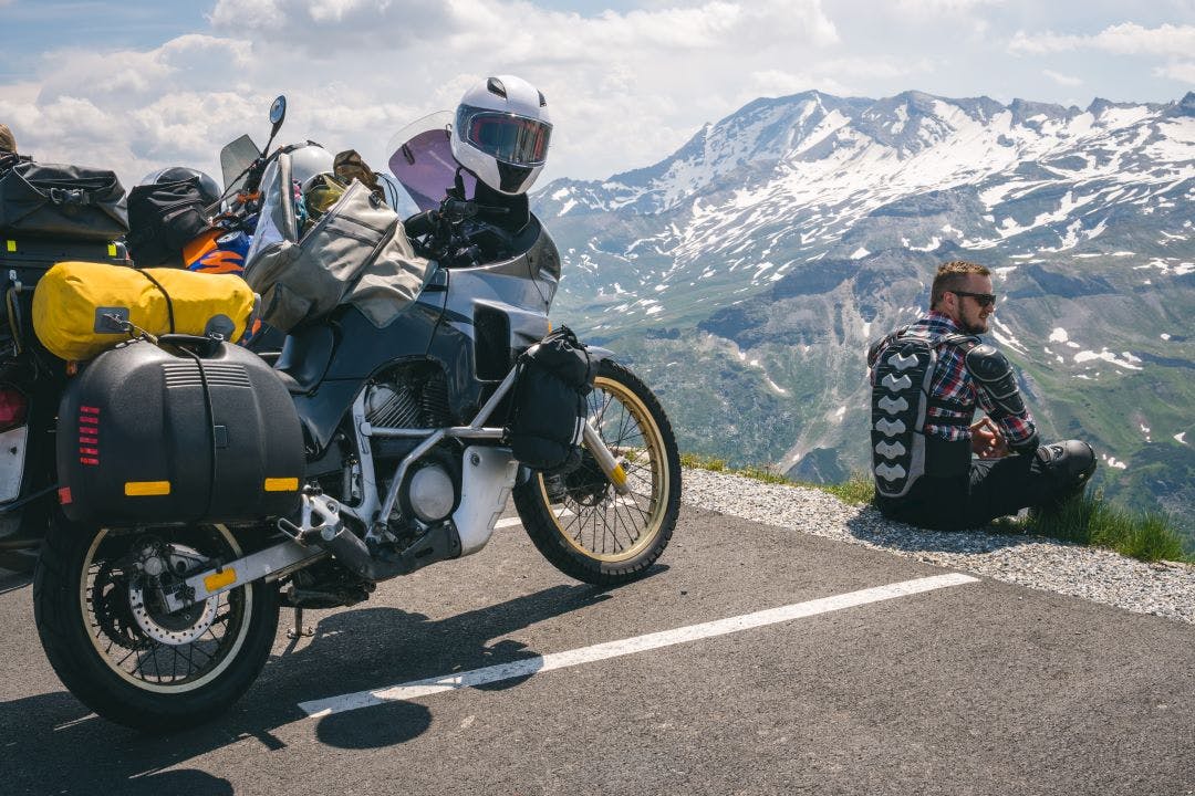 A motorcycle traveler sitting on the edge of the earth and looking into the distance. top of the mountain, Grossglockner pass, biker dressed in a protective jacket armor. What Nobody Tells You About Motorcycle Travel