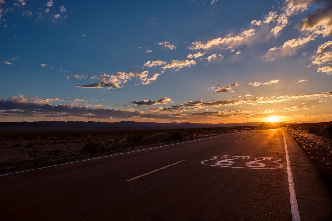 Route 66 pavement sign in the foreground and the diminishing perspective of the road leading to a dramatic sunset in the Mojave desert outside of Amboy, California 10 Most Memorable Self-Guided Motorcycle Tours in the USA